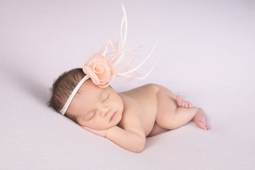Newborn rests while wearing beige headband on light pink background.