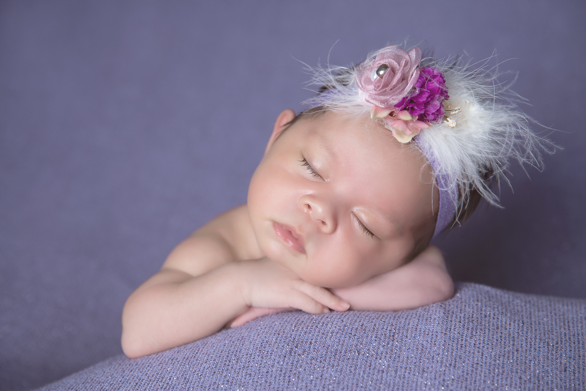 Newborn sleeps while wearing white and purple headband on purple backdrop.