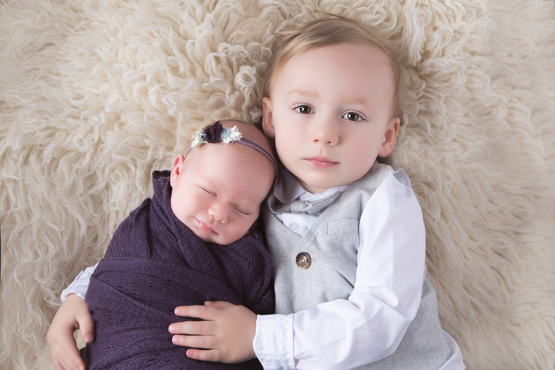 Toddler boy on gray suit holds his newborn sister which what dark purple wrap and headband. Light color carpet on background.