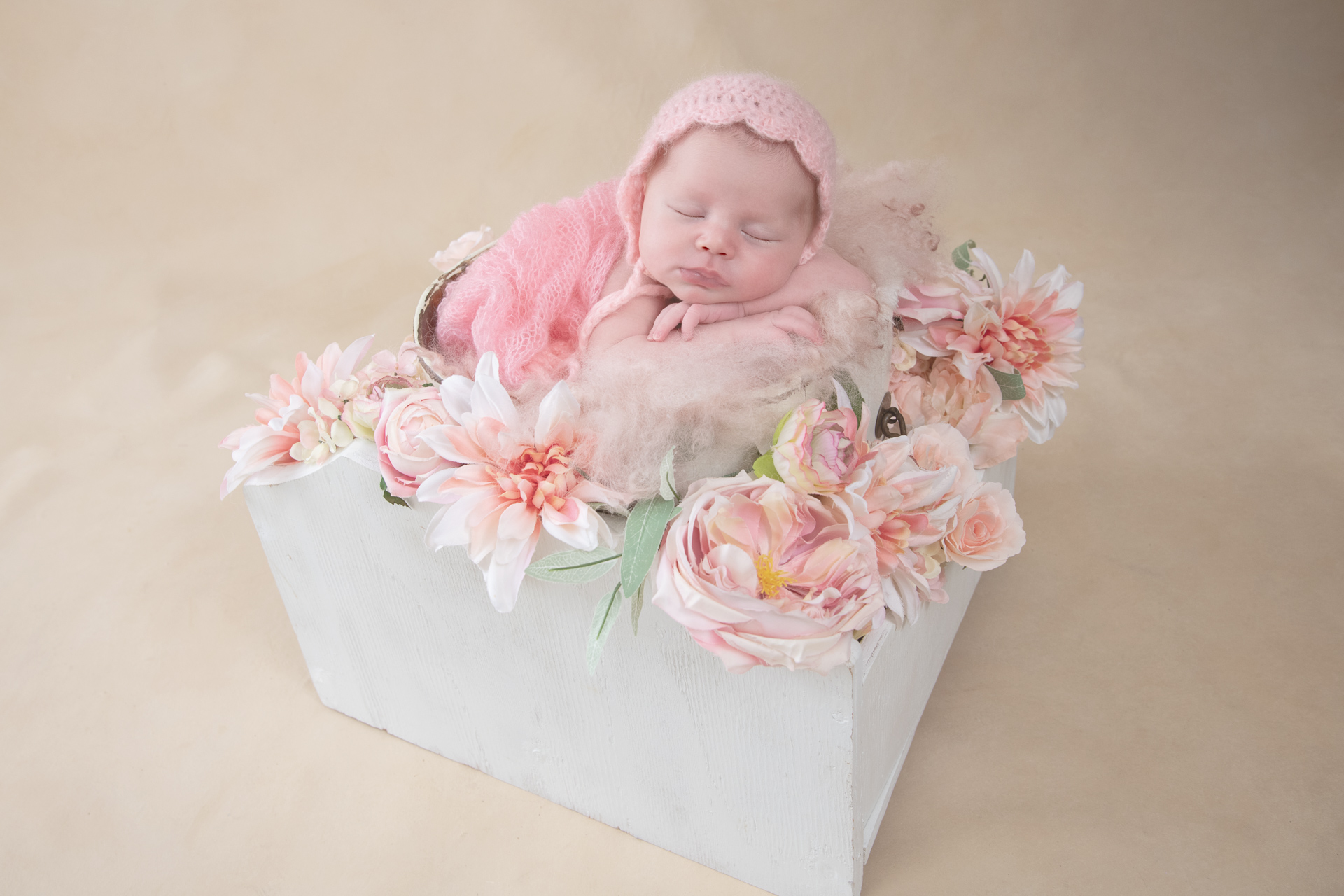 Newborn wearing pink hat and pink wrap rests on square basket prop among pink flowers which are decorating the scene. Beige backdrop.