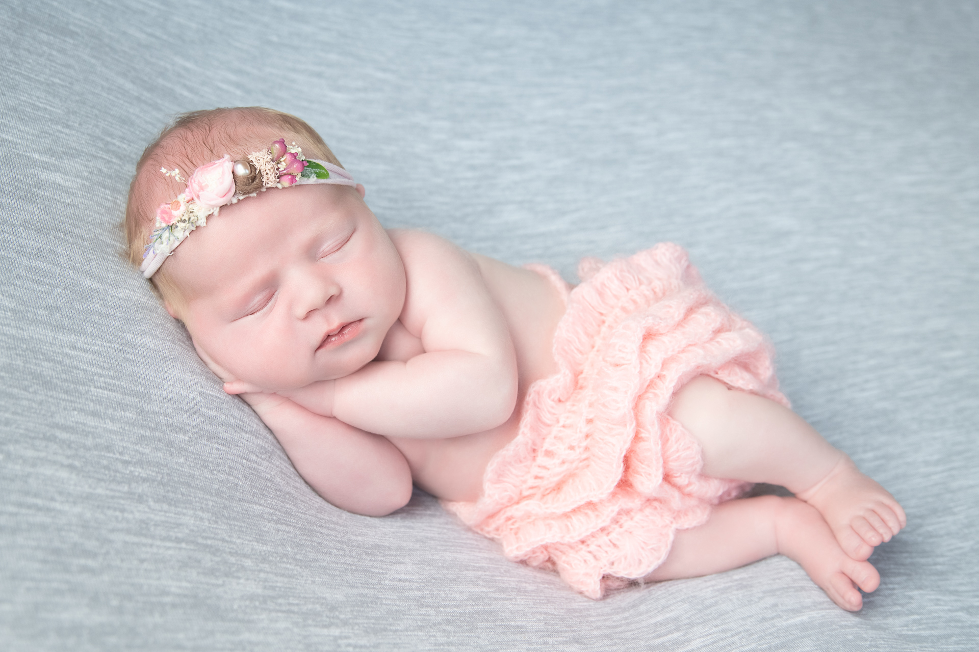 Newborn sleeps while wearing pink outfit and pink headband on light gray backdrop.