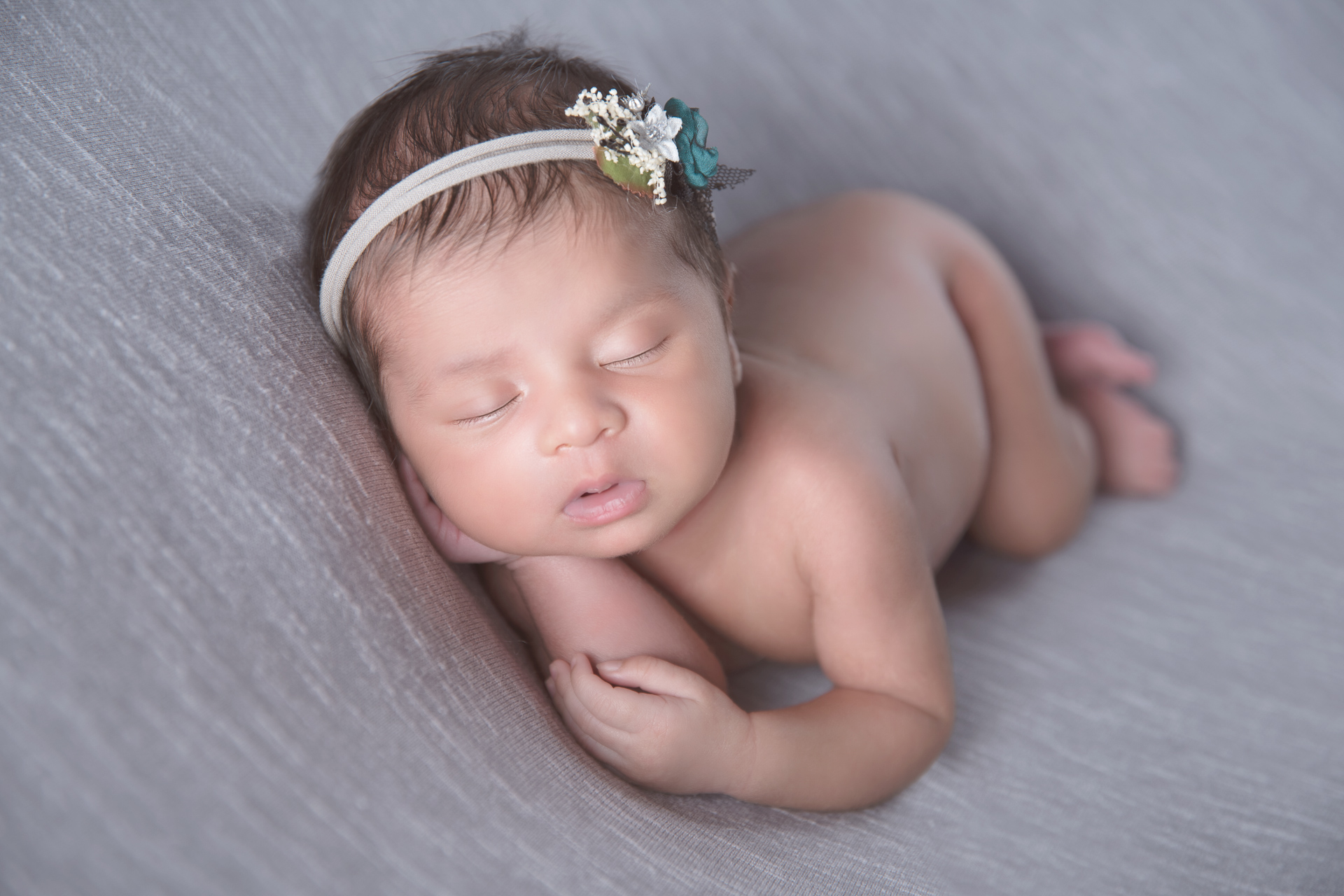 Newborn rests on gray backdrop while wearing gray headband with white and green flowers.