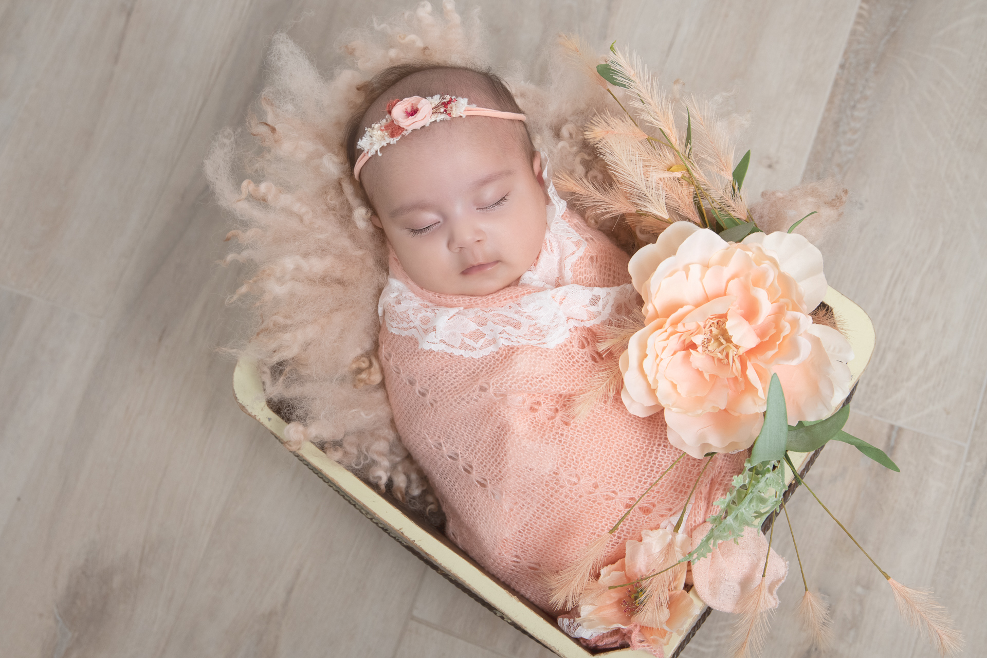 Newborn on pink headband and wrap rests on square shaped prop. Flowers and fluffy carpet decorates the scene.