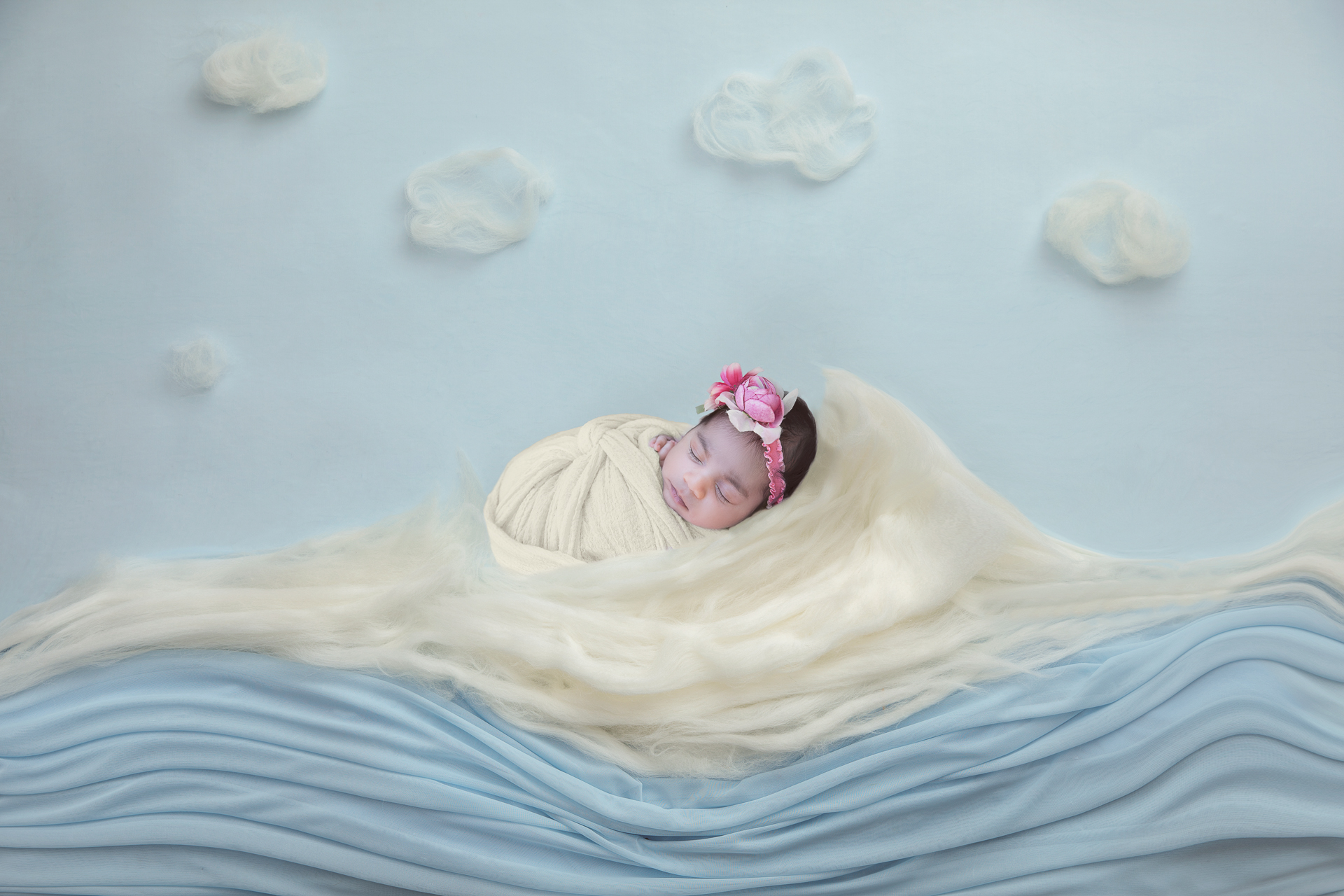 Newborn girl wearing light color wrap and pink headband appears to float on an ocean made up set up.