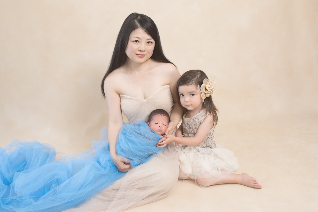 Mom wearing light brown dress indoors looks at the camera while holding her newborn baby boy next to her female older child, light brown backdrop
