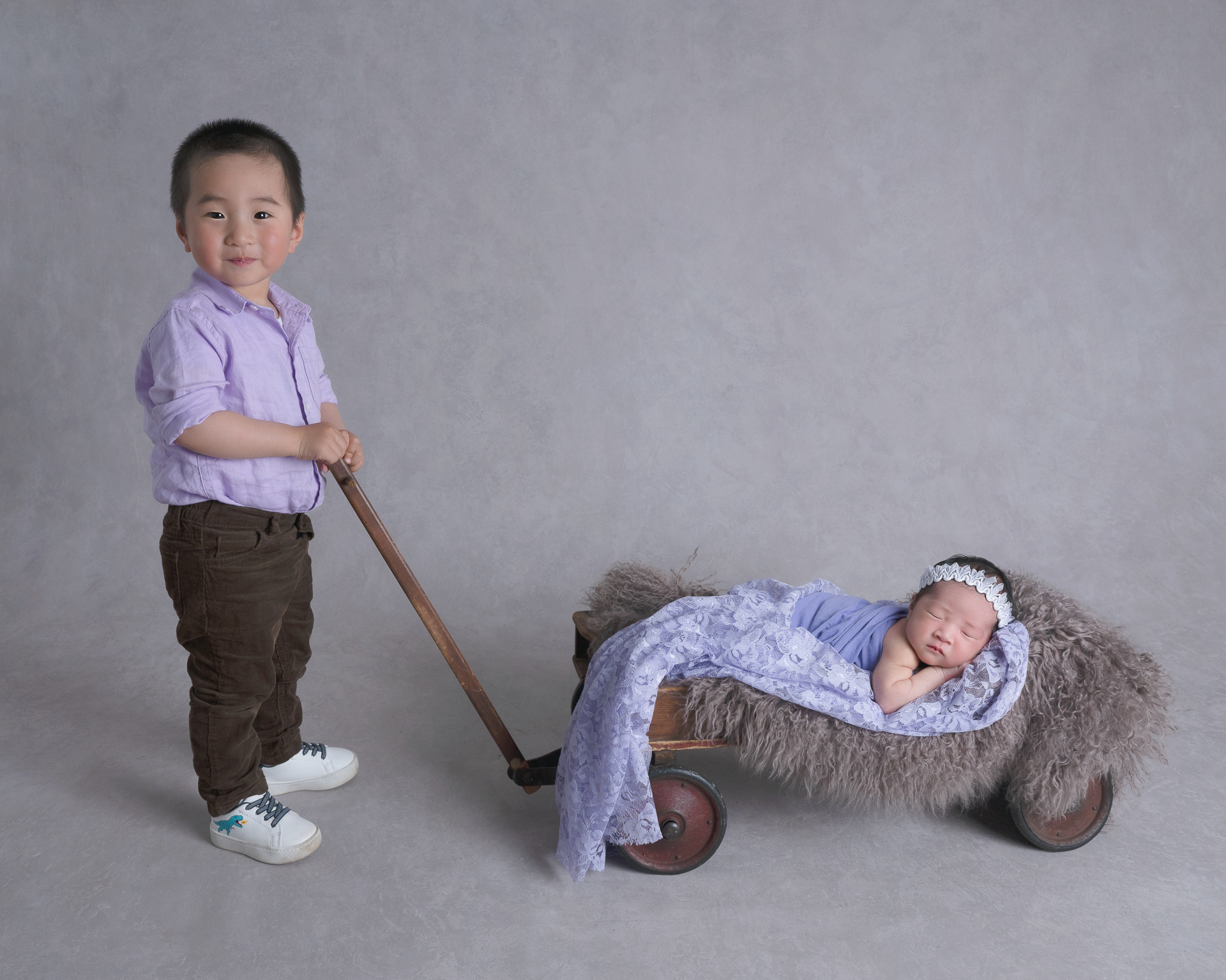Big brother holds onto wagon handle in which his newborn sister rests at. Both wear purple outfits. Baby wears light color headband. Gray backdrop.
