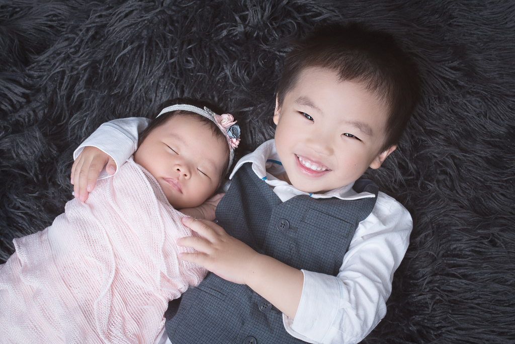 Toddler holds his newborn sister while smiling and looking at the camera. Wears dark tone suit while her sister wear pink wrap and white, pink and blue headband. Dark color fluffy carpet on the background.