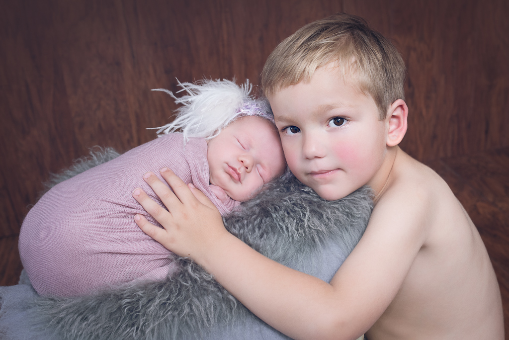 Big brother hugs newborn sister while she rest on a newborn prop. Brown backdrop, gray fluffy carpet decorates the scene.