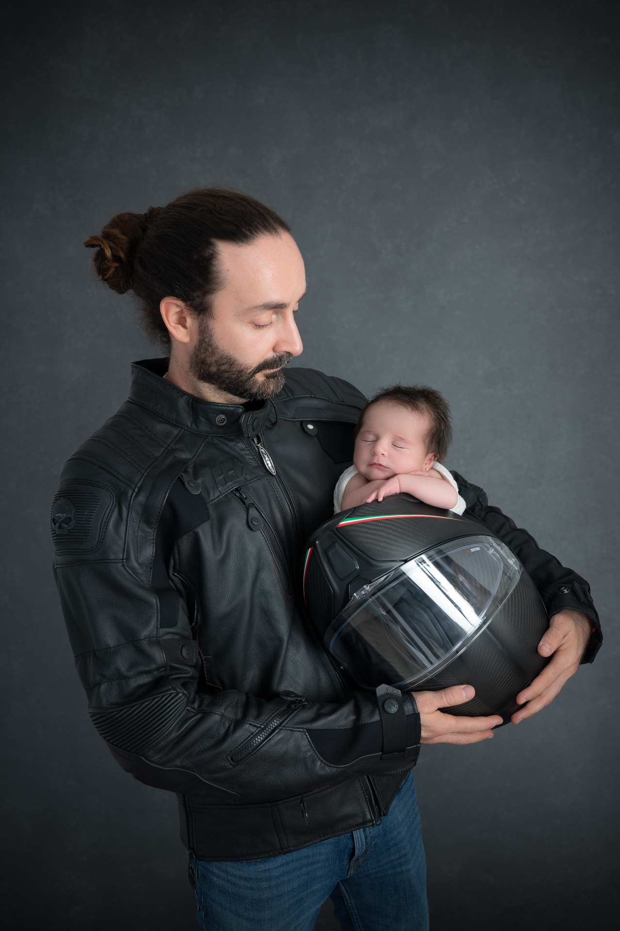 Man holds his newborn daughter which is resting on motorcycle helmet. Wears motorcycle black leather jacket and blue jeans. Dark backdrop.