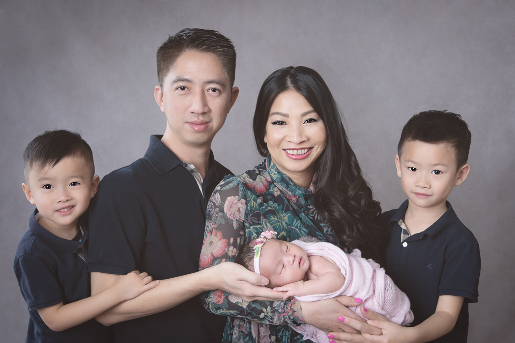Family of 4 poses indoors on gray backdrop. Newborn girl wears pink headband and pink wrap. Mom wears flowers dress, dad and older kids on dark T-shirts.