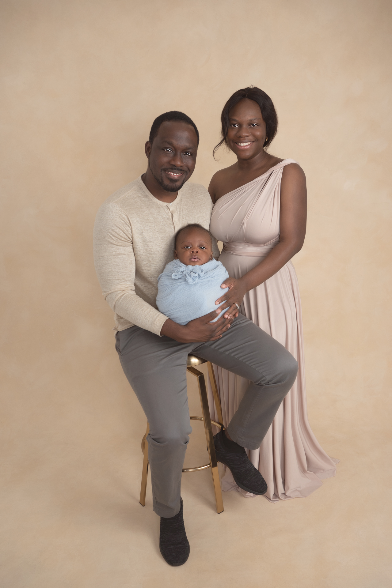 Couple posing indoors with their newborn son. Light brown outfits. Baby wears light blue wrap. Light brown backdrop.