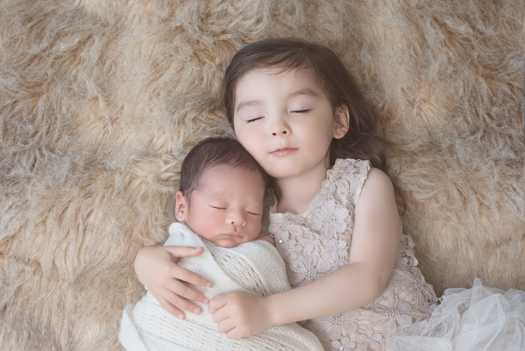 Toddler sister hugs her newborn brother. Brown tones outfits. Brown carpet