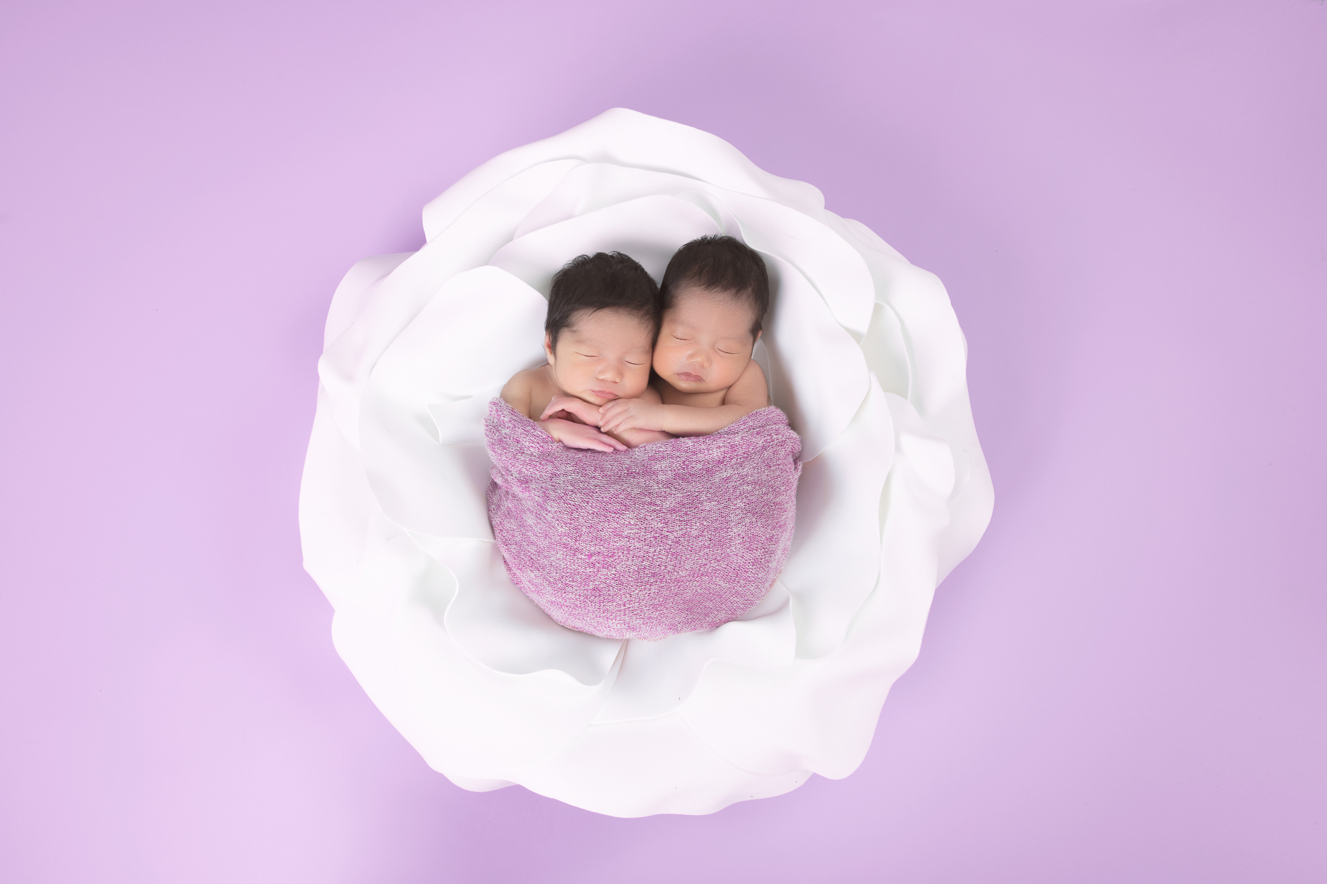 2 newborn sisters rest together on a white prop, wearing pink wrap, on a pink backdrop