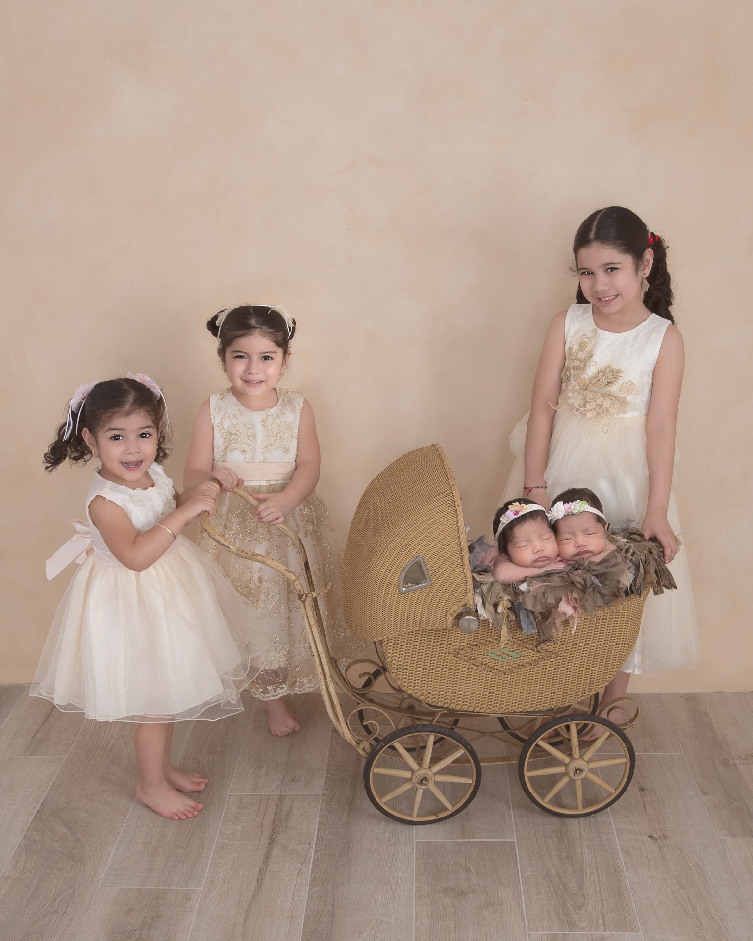 3 girls posing with their newborn twin sisters that are resting on a old fashioned decorative stroller. Light brown backdrop.