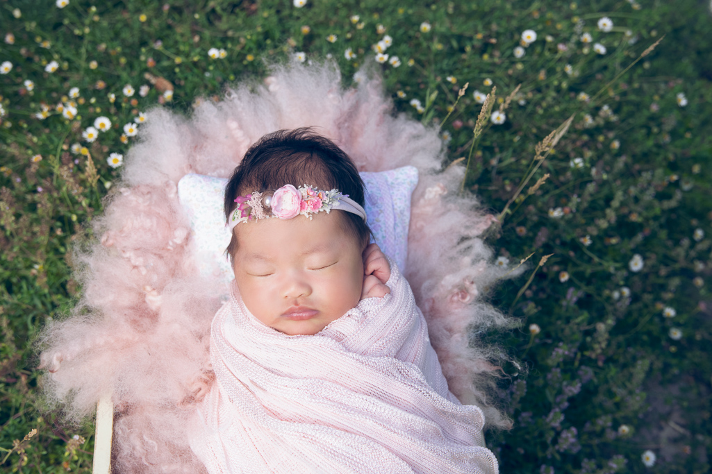 Newborn girl rests on a prop outdoors wearing pink headband and pink wrap. Blurry flower margaritas and grass decorating the scene.