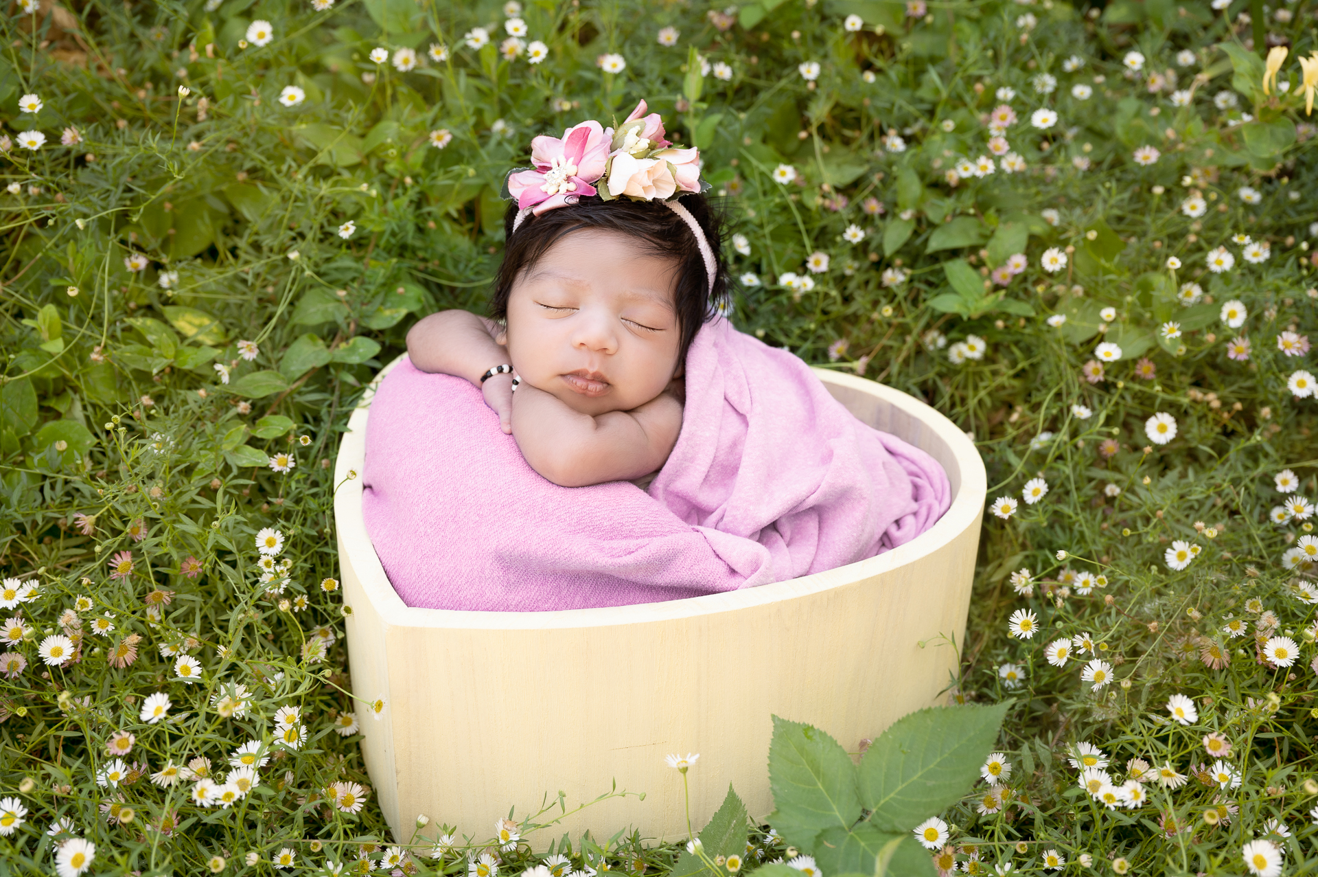 Newborn girl rests on heart shaped prop outdoors surrounded by margarita flowers. Pink blanket & headband.