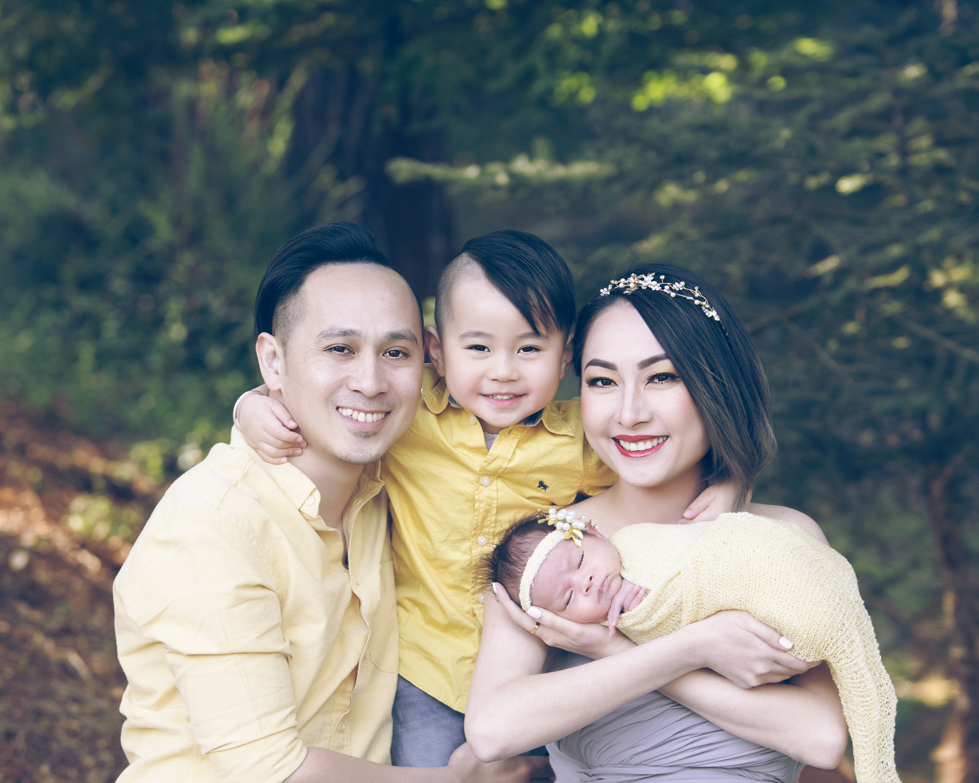 Family of 4 posing outdoors. Father on yellow T-shirt, toddler boy on T-shit, mother on gray dress holding newborn baby on yellow wrap outfit.