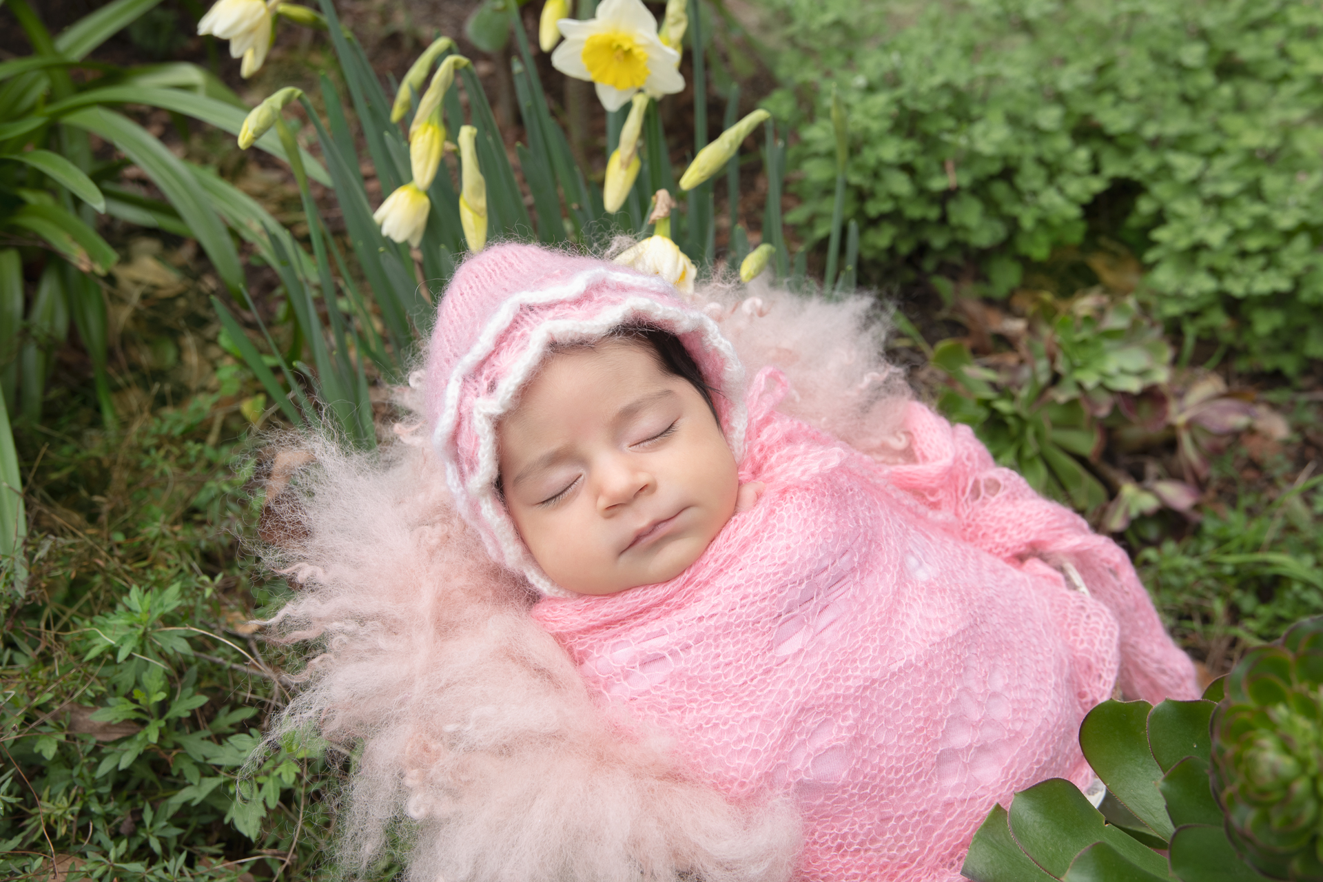 Newborn girl rests outdoors on pink outfit and pink prop. Pink hat. Flowers and grass decorates the scene.