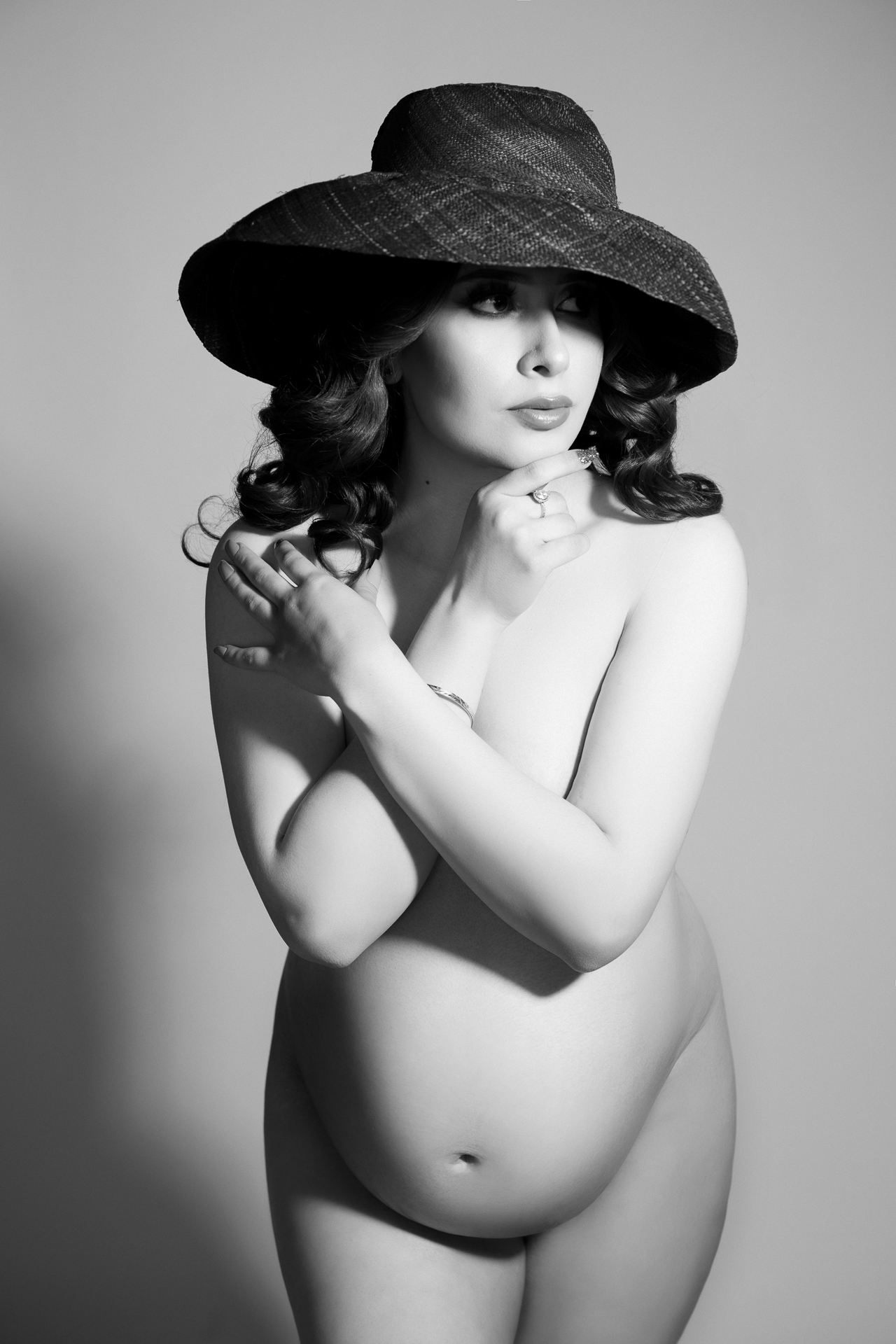 black and white image, non dress pregnant woman wears dark hat on white backdrop