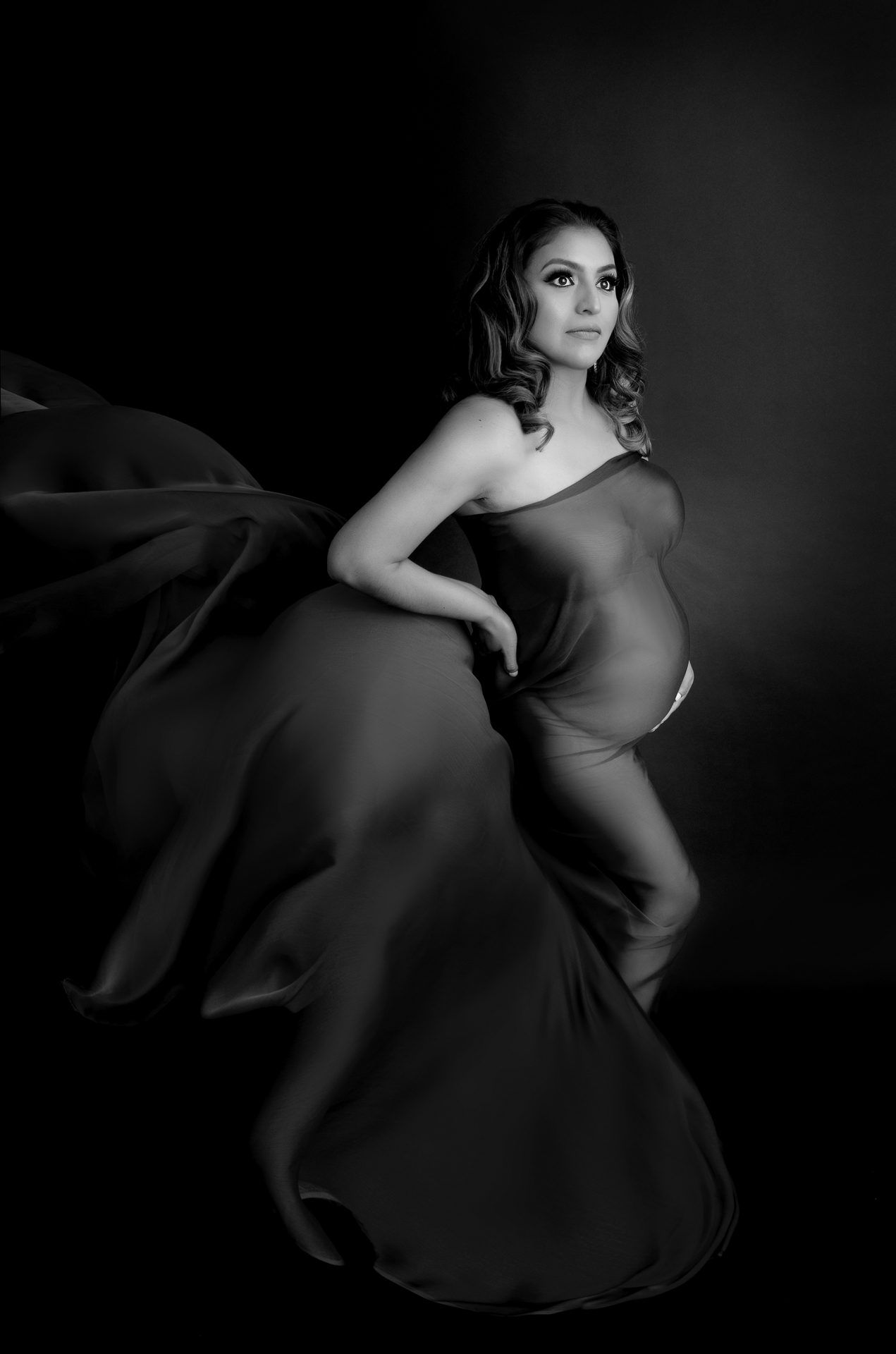 Pregnant woman wears dar fabric dress, dark backdrop, black and white image.