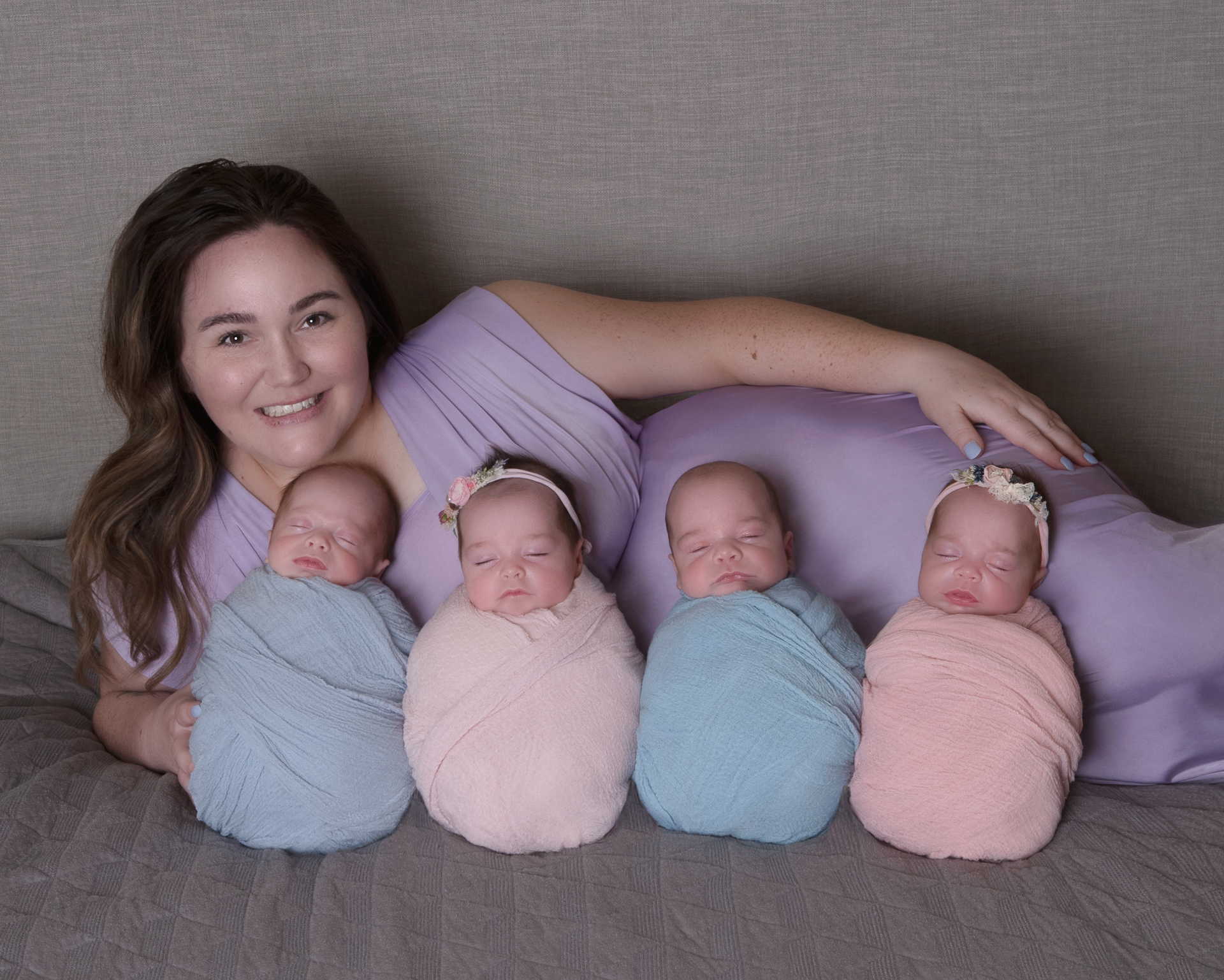 Mother wearing purple dress lays on a bed posing indoors with her four newborns which are placed in front of her. Two of them wear pink wrap outfits and headbands, two of them wear blue wrap outfits.