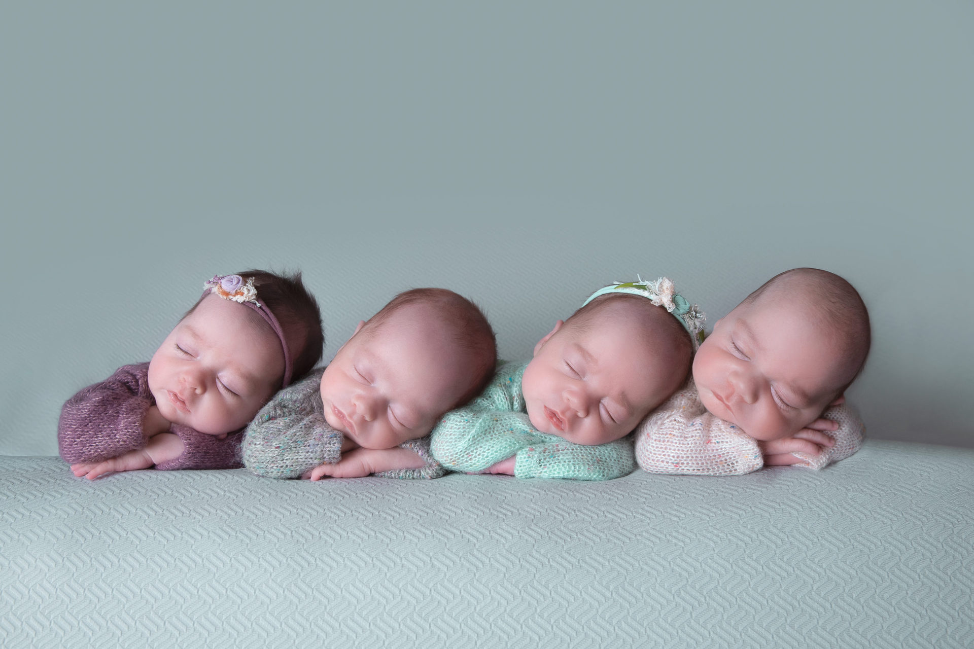 4 newborn siblings posing indoors on light green backdrop. Straight on angle shot. Girls wear purple and green outfits and headbands, while boys wear gray and light colored outfits.
