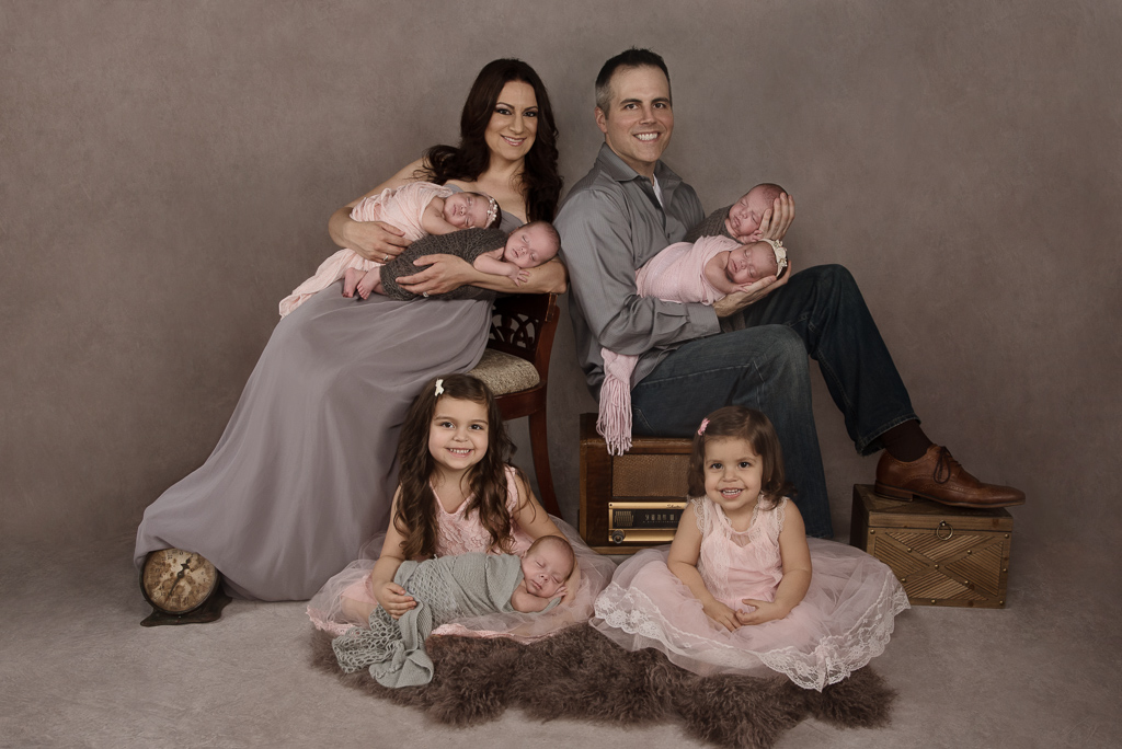 Family of 7 poses indoors. Mom and dad, 5 newborn girls and two 4 year old girls.