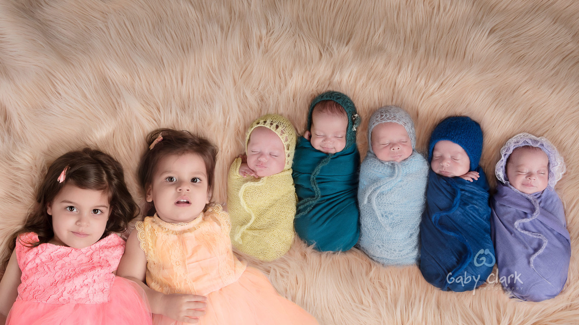 90 degrees shot shows 5 newborn siblings and their older sisters. Light brown fluffy carpet background. One different color is assigned to each child. Pink, yellow, yellow, green, light blue, dark blue & purple.