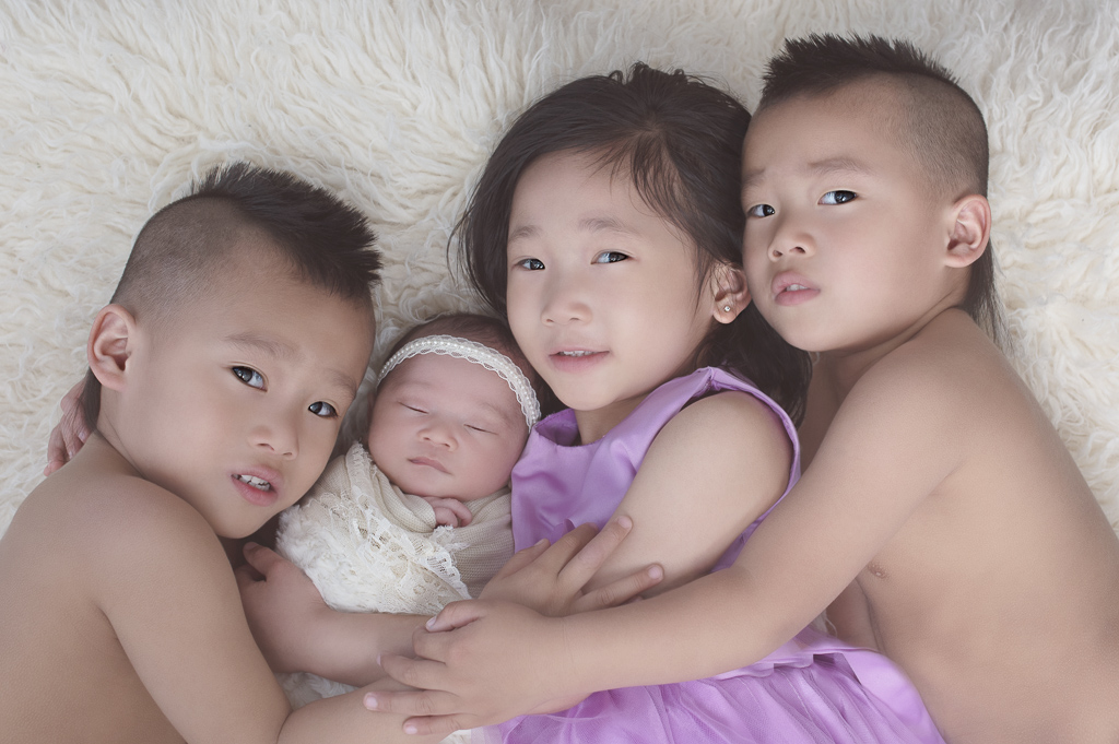 3 older siblings hug their newborn sister. Baby wears light color headband and light color wrap. Light color fluffy carpet on the background.