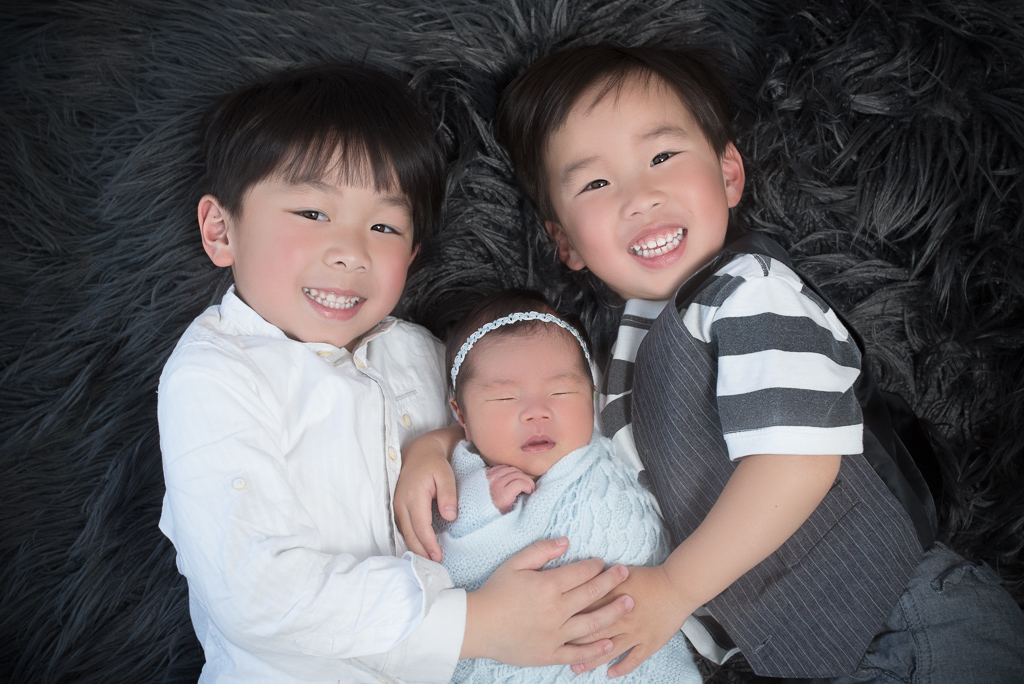 Two toddlers siblings hold their newborn sister on dark carpet background