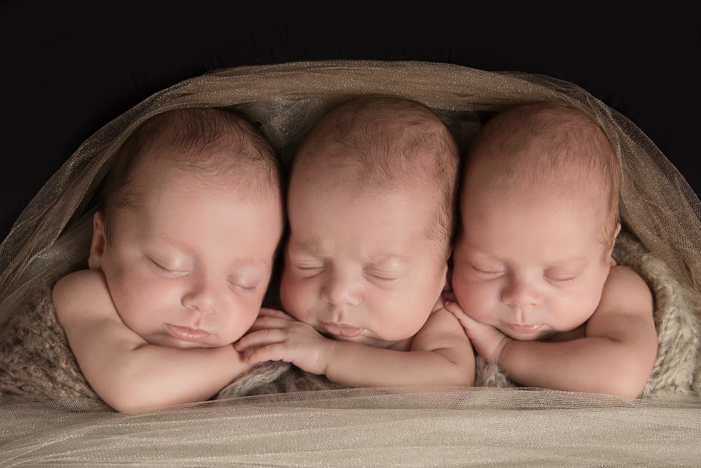 3 newborn siblings rest together wrapped on light brown fabrics tones