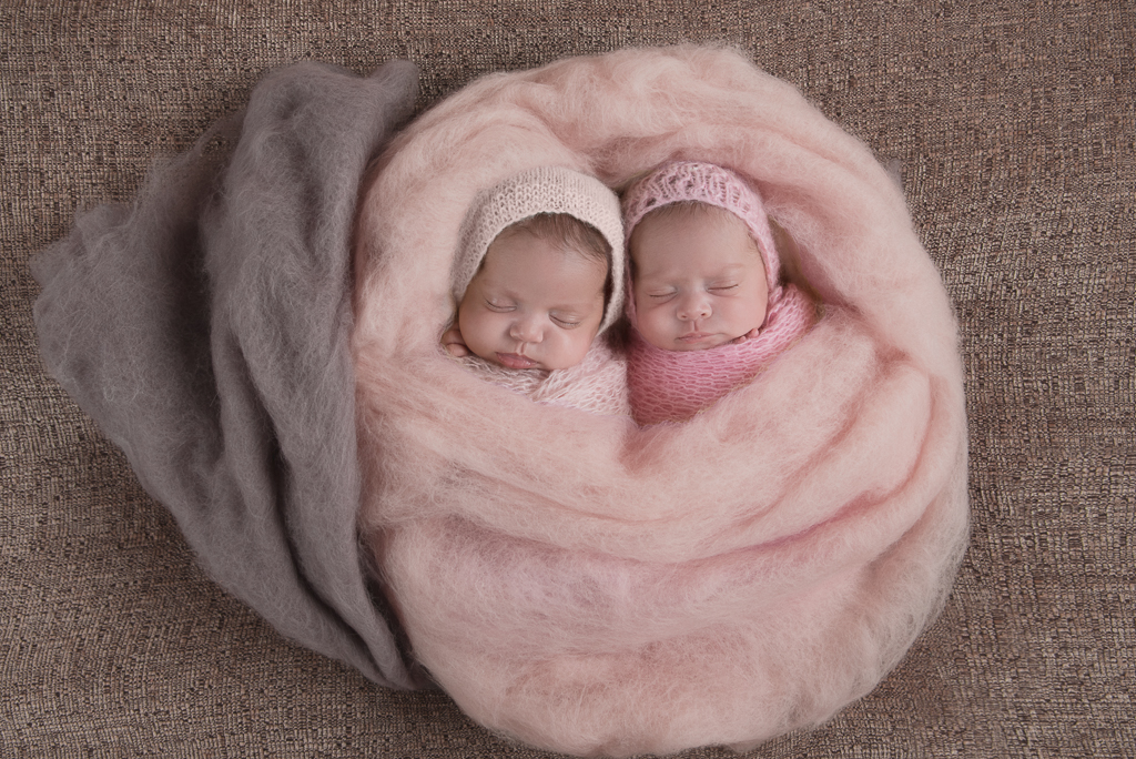 2 newborn babies rest together wearing pink tone wraps and hats. pink decorative carpet wrap them up as well.