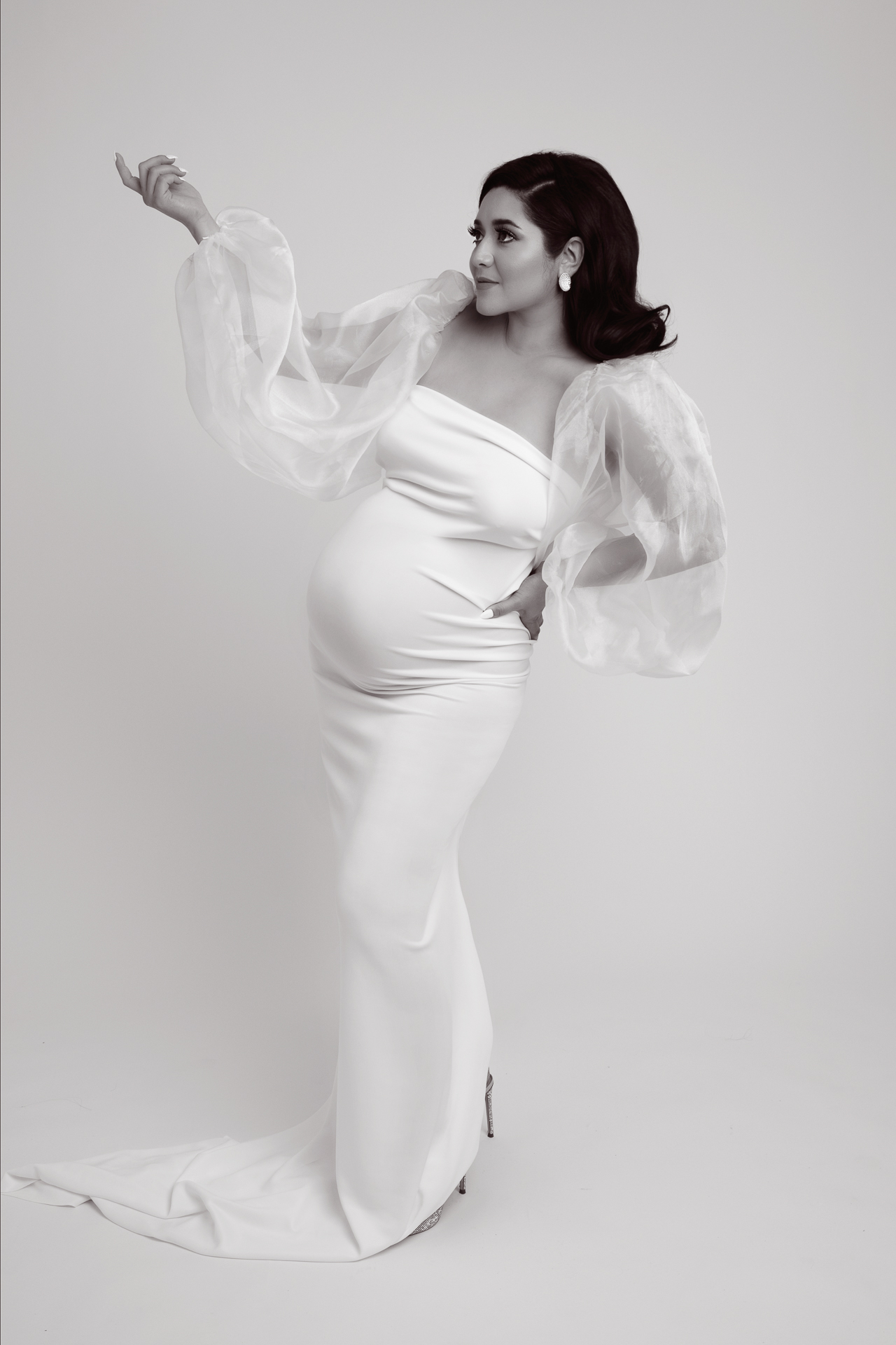 Pregnant, dark hair woman wears white dress and high heels, white backdrop, black and white image