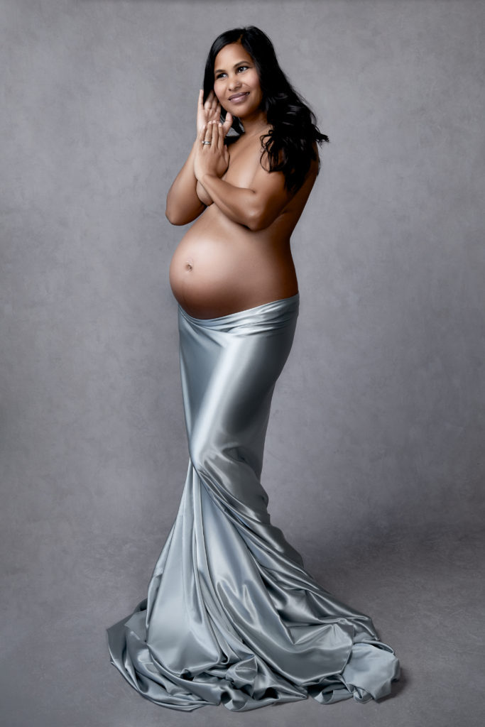 Pregnant woman during her maternity photo shoot wears shiny light gray fabric. Gray backdrop.
