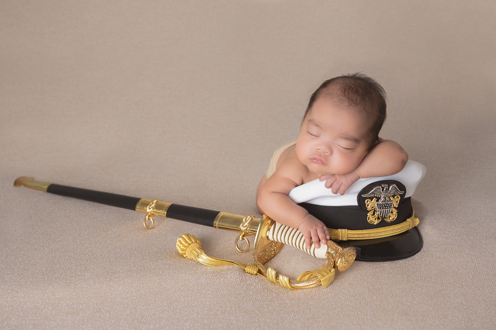 newborn baby boy resting on top of his fathers marine hat while holding onto his fathers marine sword after retouching