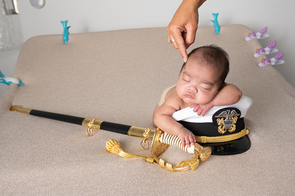 newborn baby boy resting on top of his fathers marine hat while holding onto his fathers marine sword before retouching