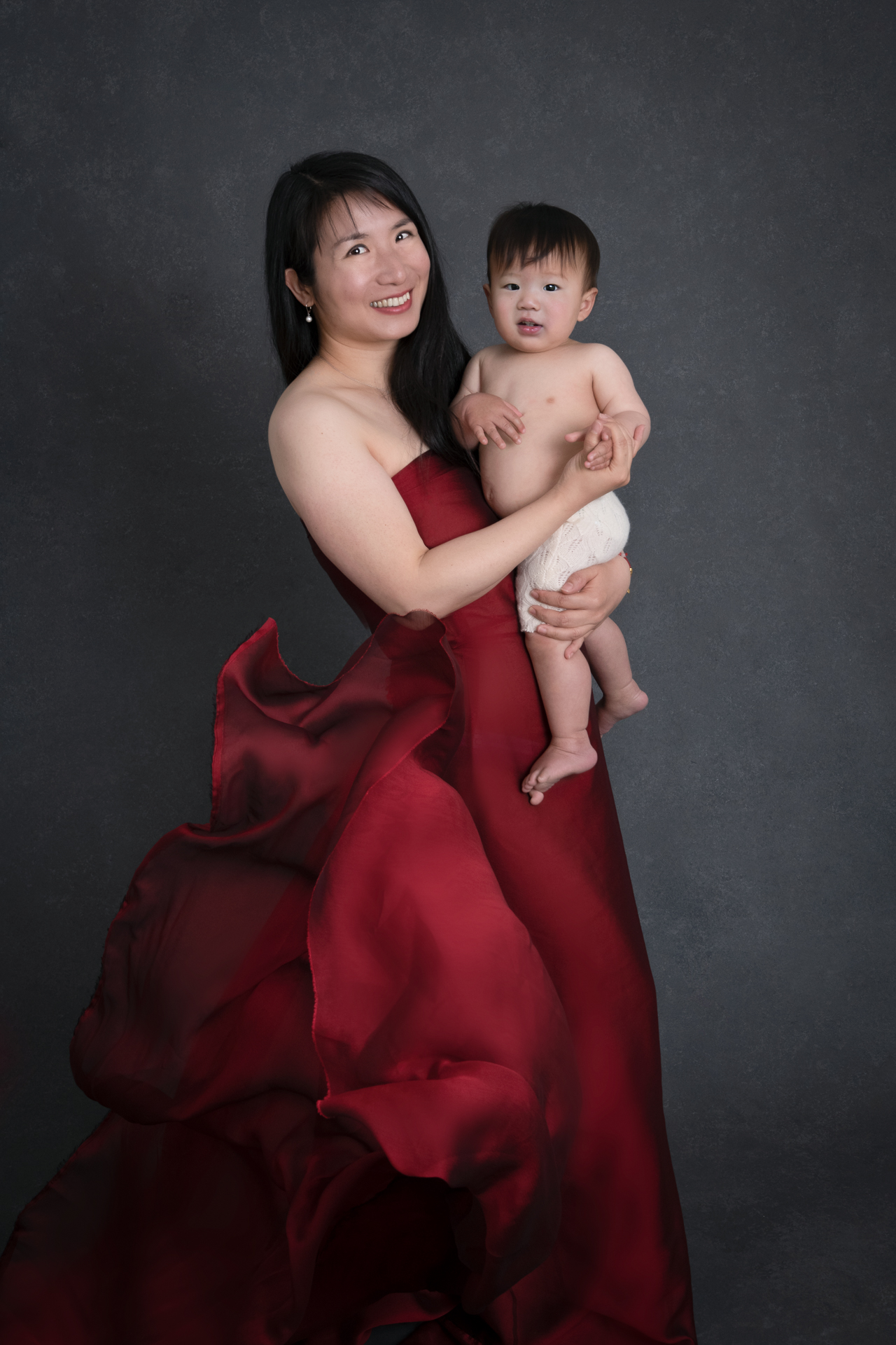 mother holding her one year old son while wearing red dress, dark gray backdrop
