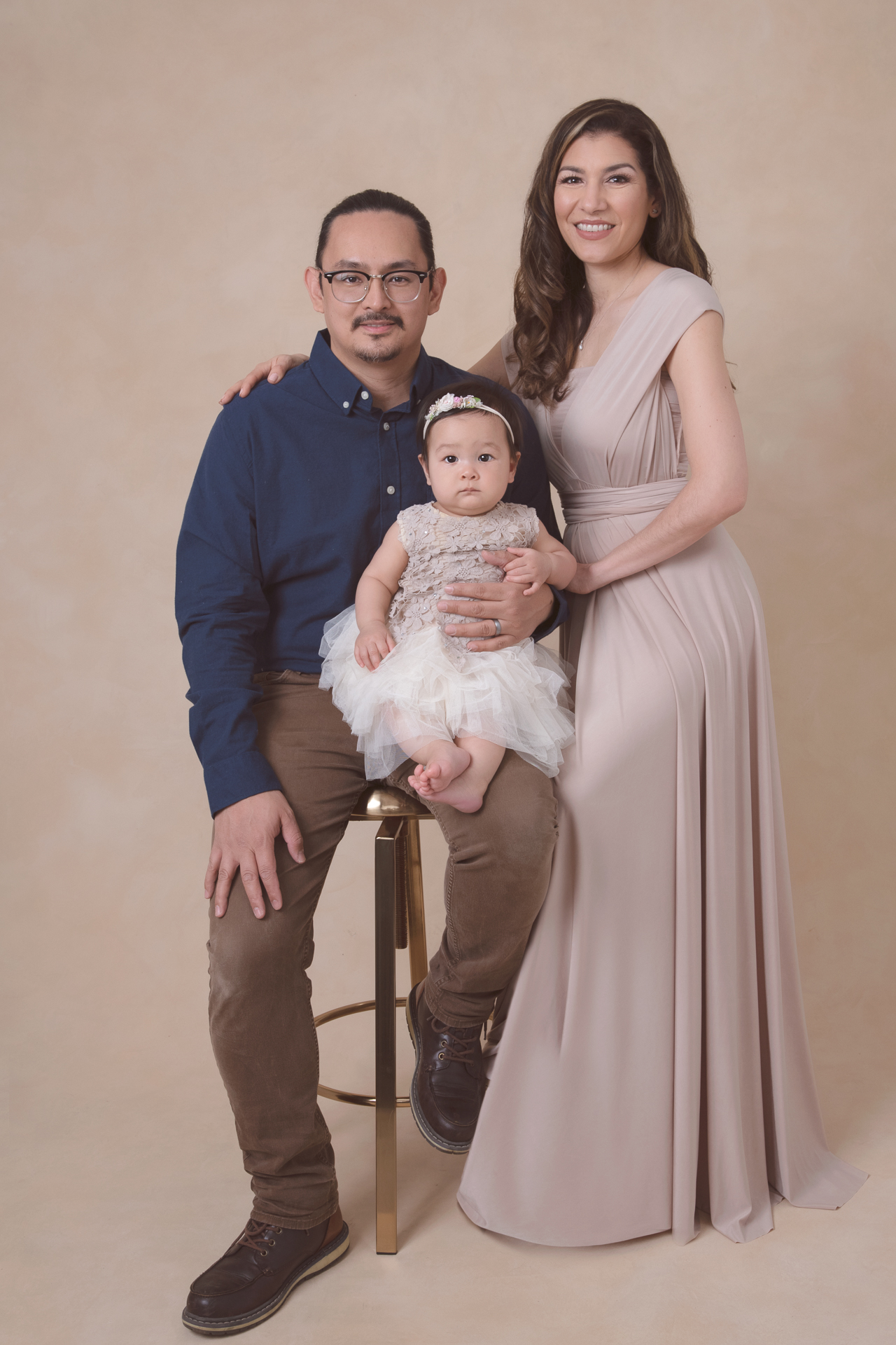 mother and father posing for the camera while holding their one year old girl, mom wears beige dress, baby wears beige color dress and dad wears a blu T shit, beige backdrop