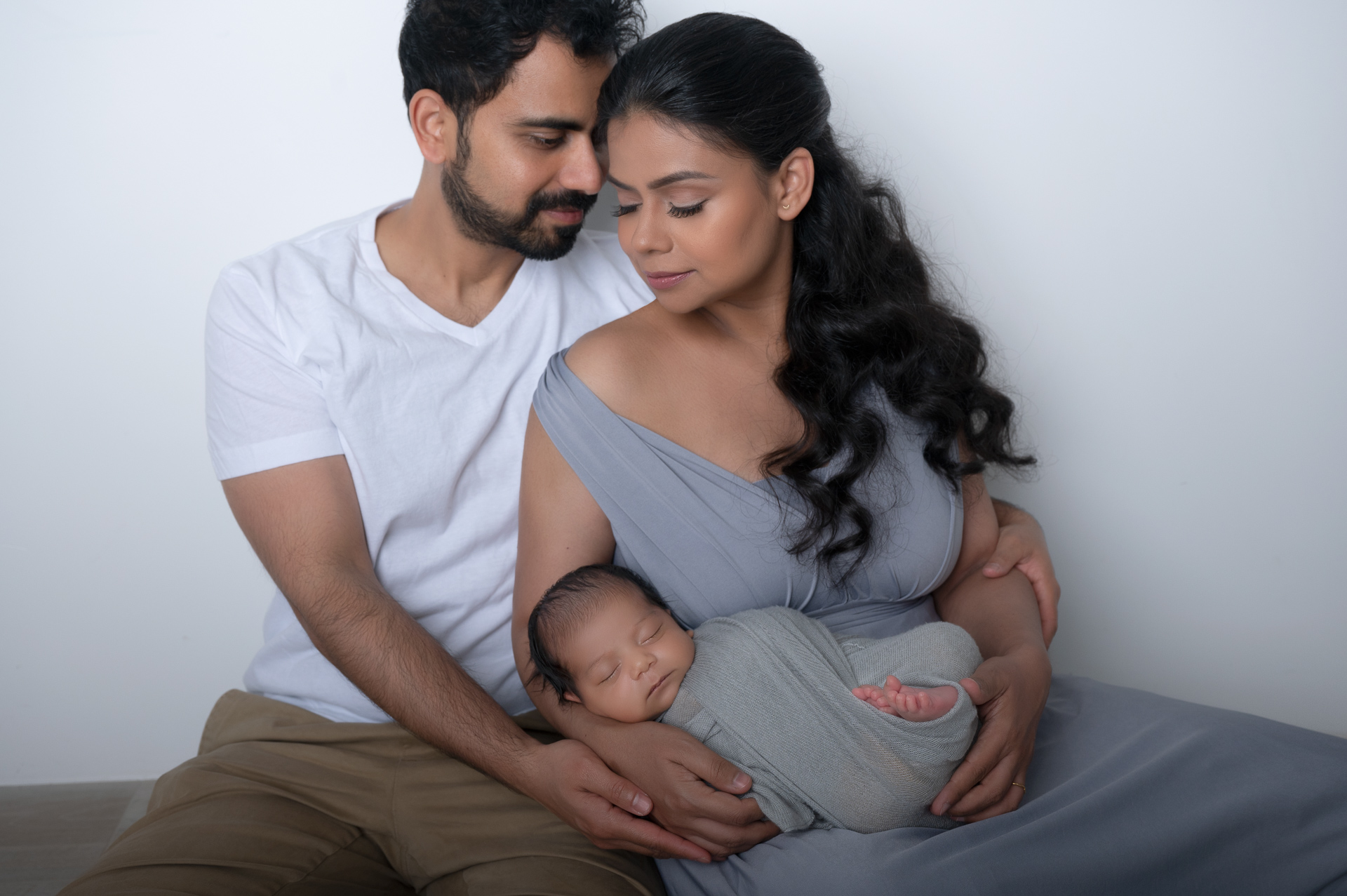 mom and dad holding their newborn son sitting and look away from the camera, white backdrop