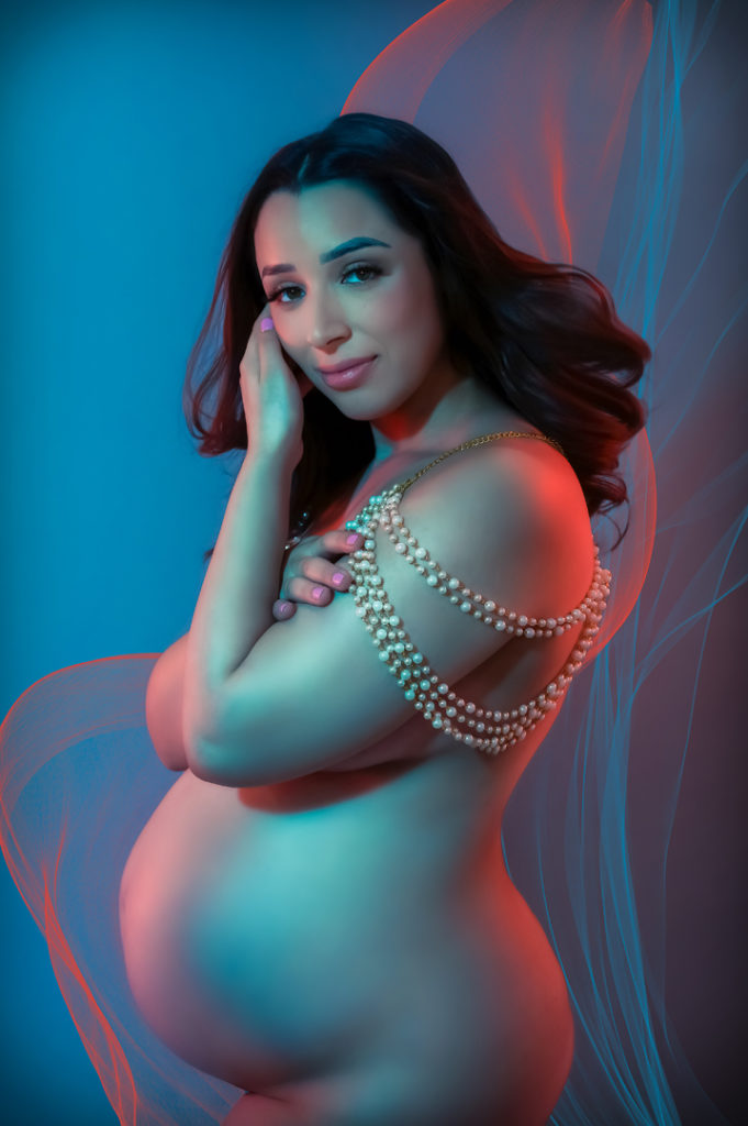 pregnant woman wearing necklace looks at the camera while illuminated by orange and turquoise colorful gel lighting