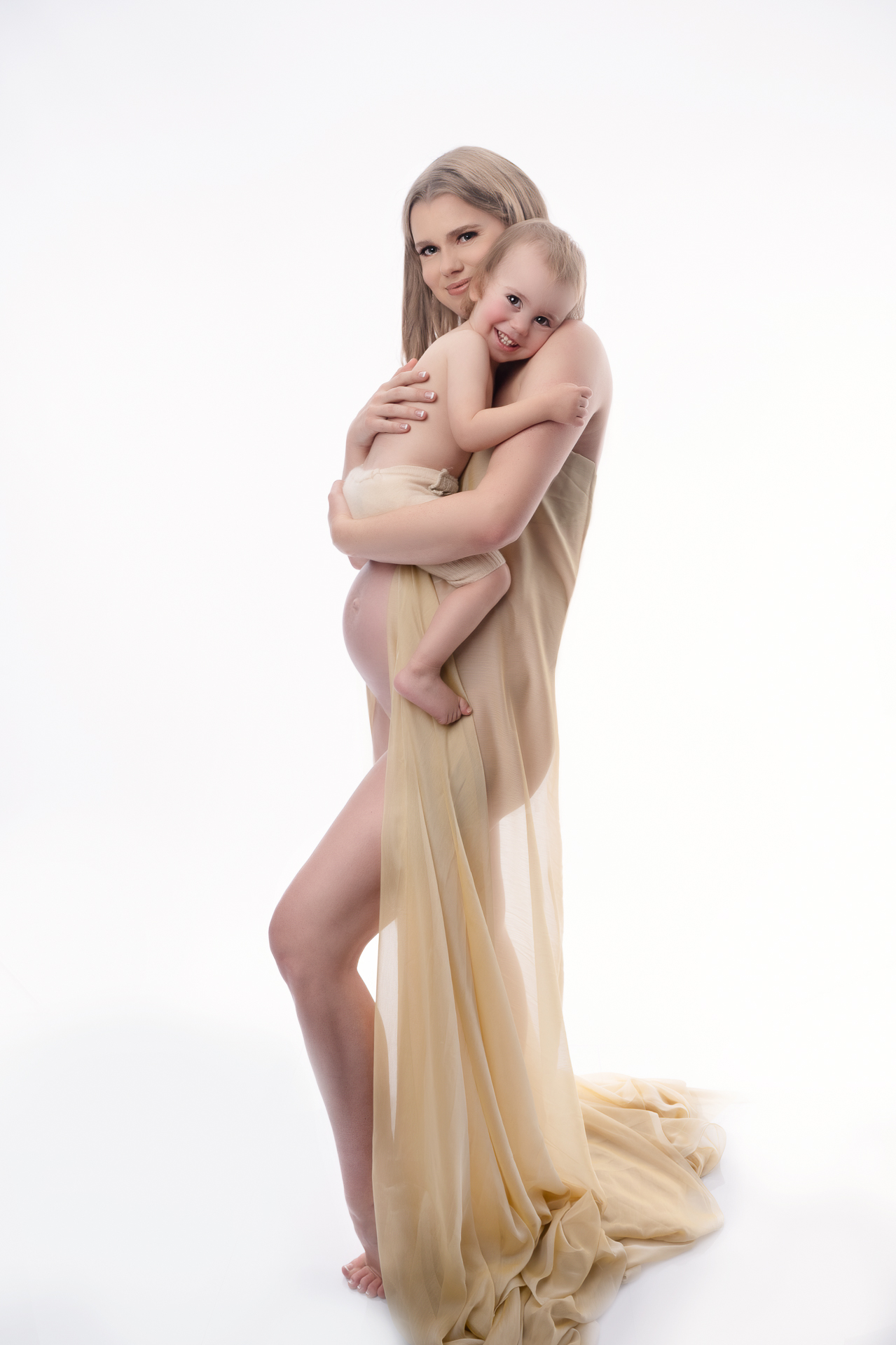 pregnant woman holds her toddler son while wearing a golden fabric dress, white backdrop
