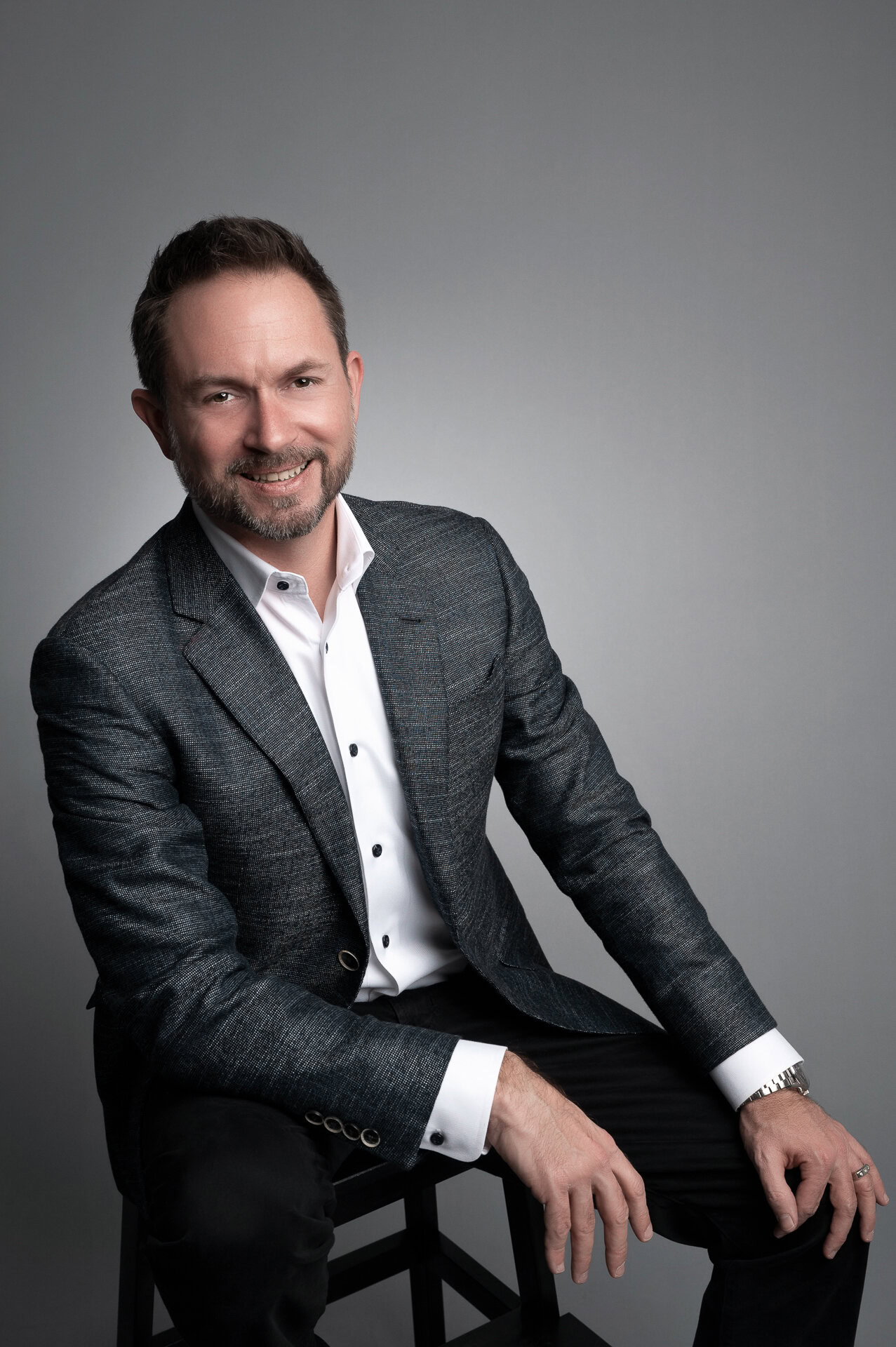 Professional Headshot of a Man in a Suit A smiling man in a gray blazer sits on a stool against a gray backdrop, perfect for personal branding photography session.