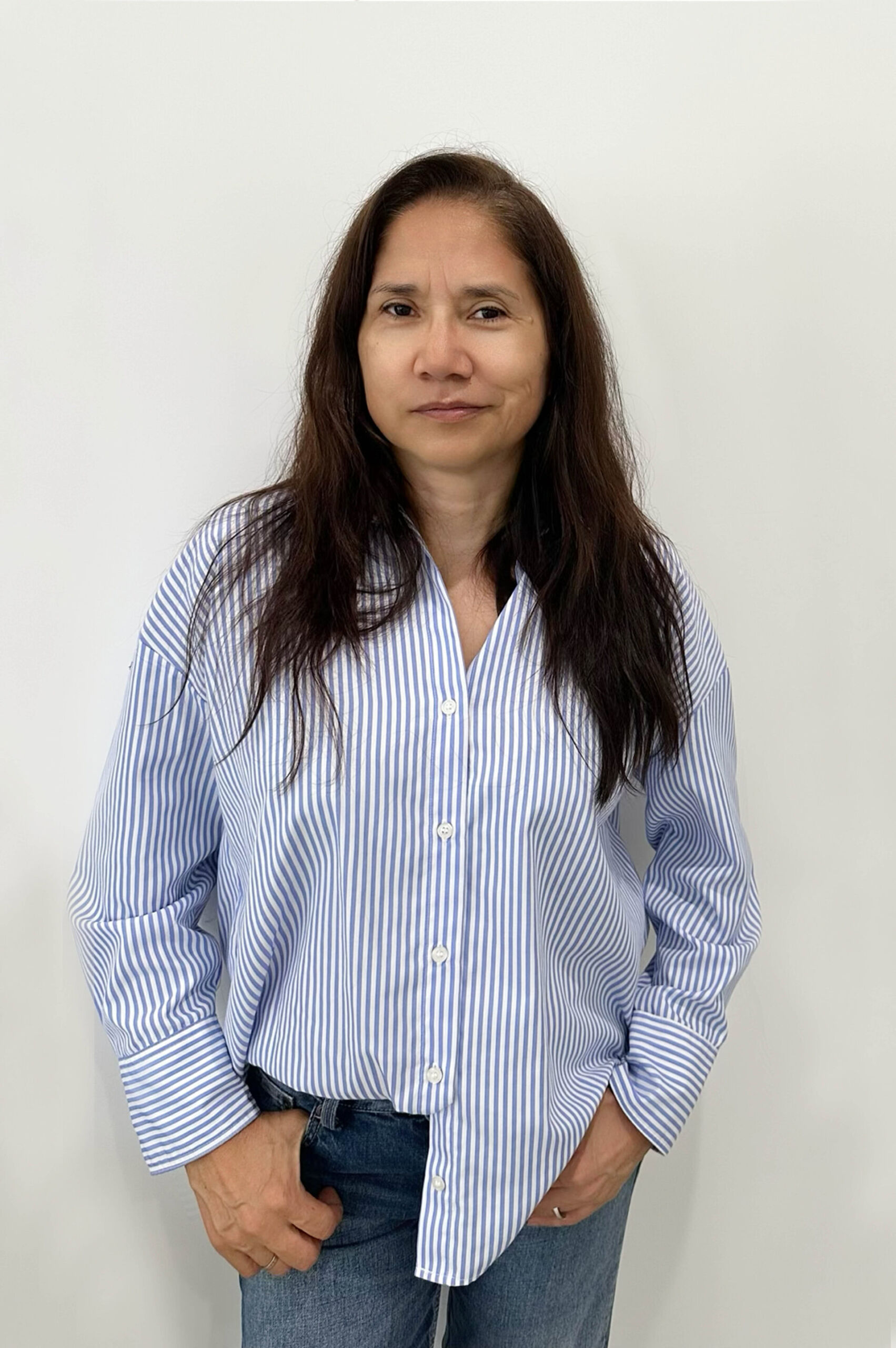 Woman with long dark hair wearing a blue and white striped shirt and jeans, standing against a plain background.