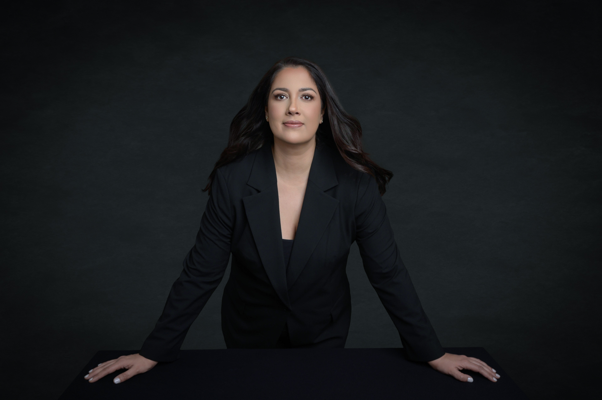 Personal branding: A confident person with long dark hair in a black suit stands behind a table, gazing upward against a dark backdrop.
