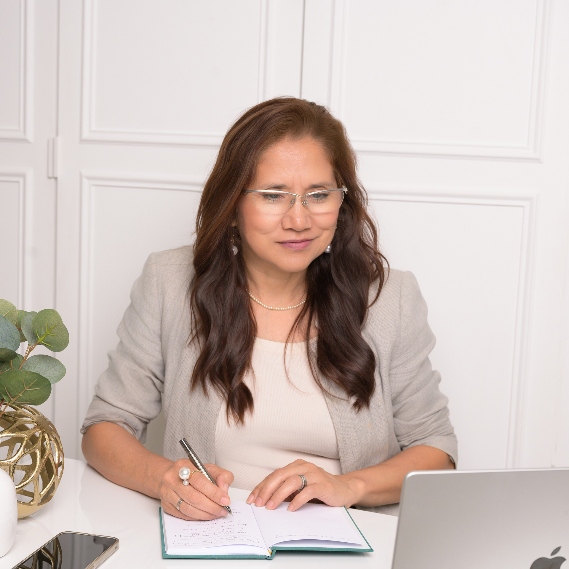 Personal branding: A woman with long brown hair and glasses works at a desk with a laptop, notebook, plant, and decor nearby.