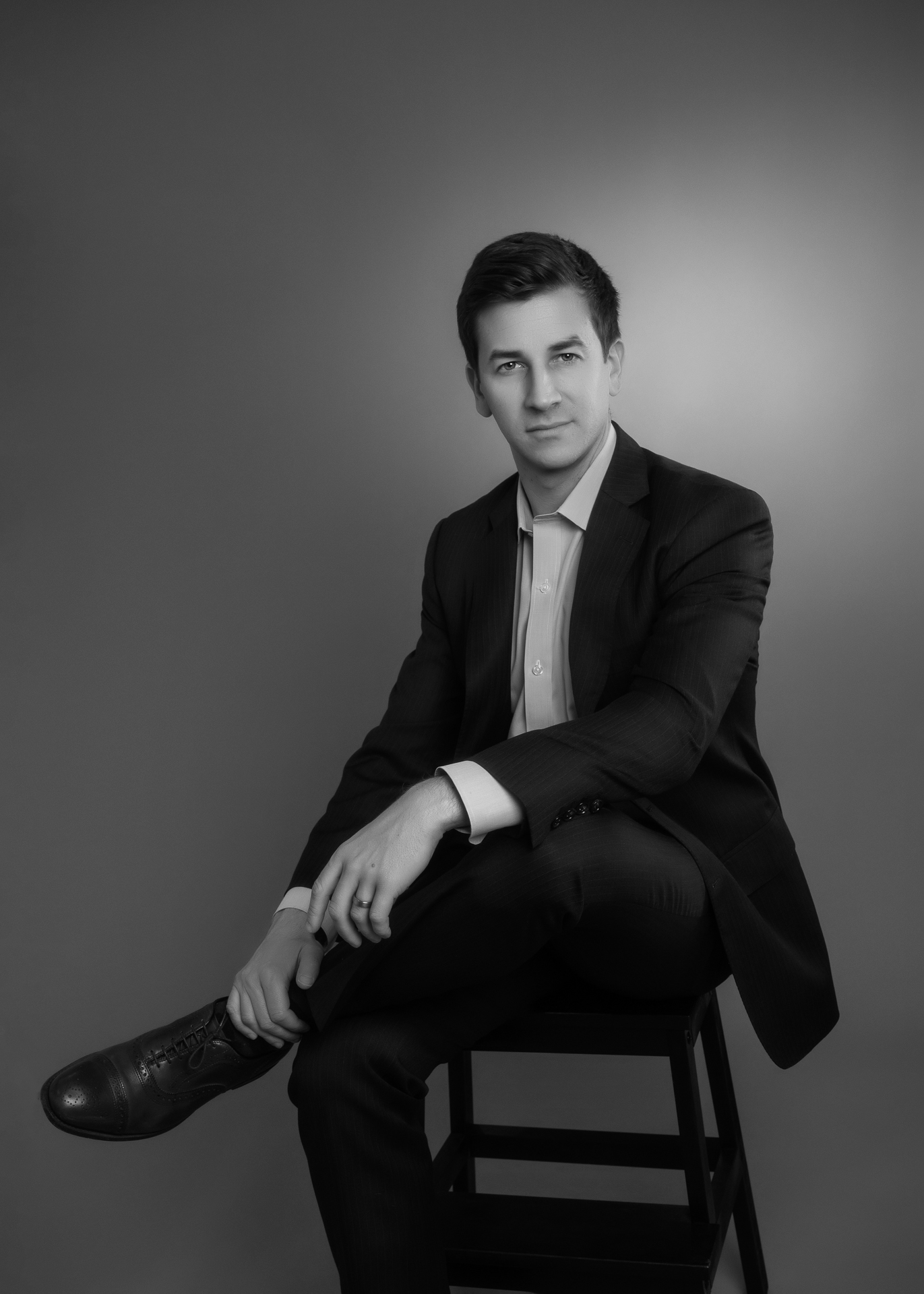 headshot portrait of a man in a suit sitting on a stool, facing the camera, in a black and white studio photo.
