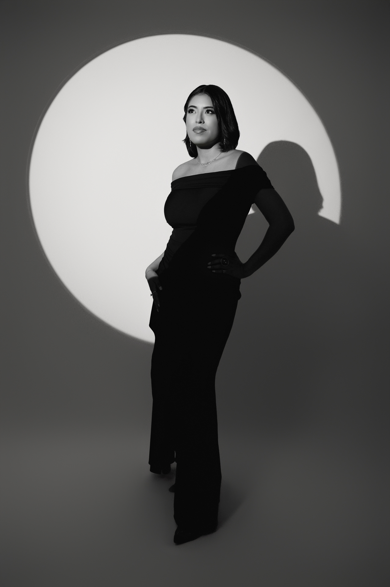 Personal branding photo of a woman in an elegant black dress, posing confidently with dramatic lighting and shadows in a monochrome studio.