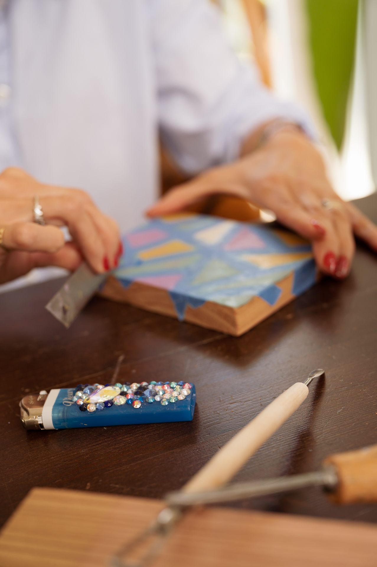 Image by Gaby Clark www.gabyclark.com A person cuts blue painter’s tape on a painted wood table; beads, a lighter, and a metal tool sit nearby. No people shown.