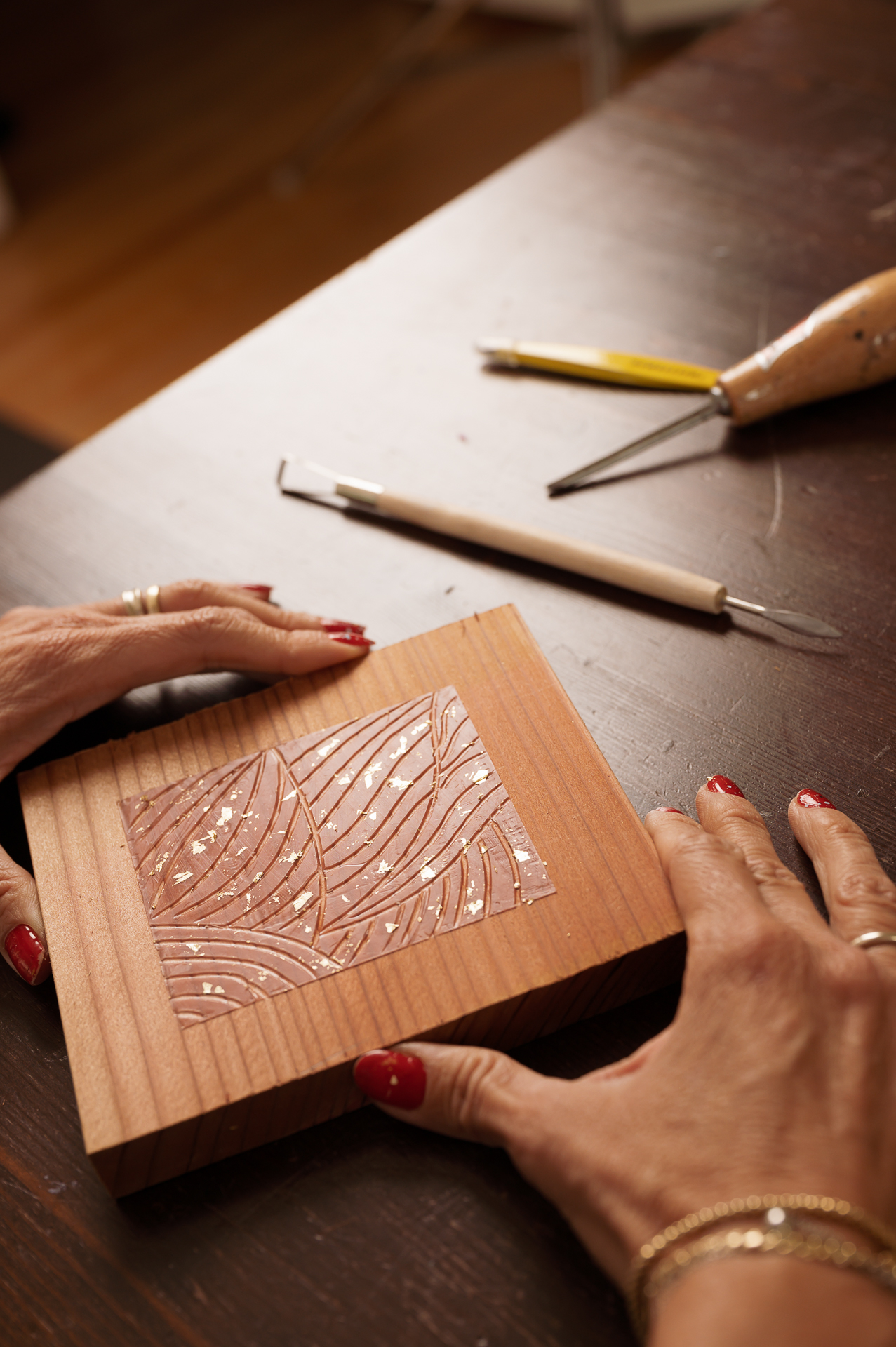 Image by Gaby Clark www.gabyclark.com Personal branding: A hand with red nails holds a carved wooden block; two carving tools rest on a dark wooden table.