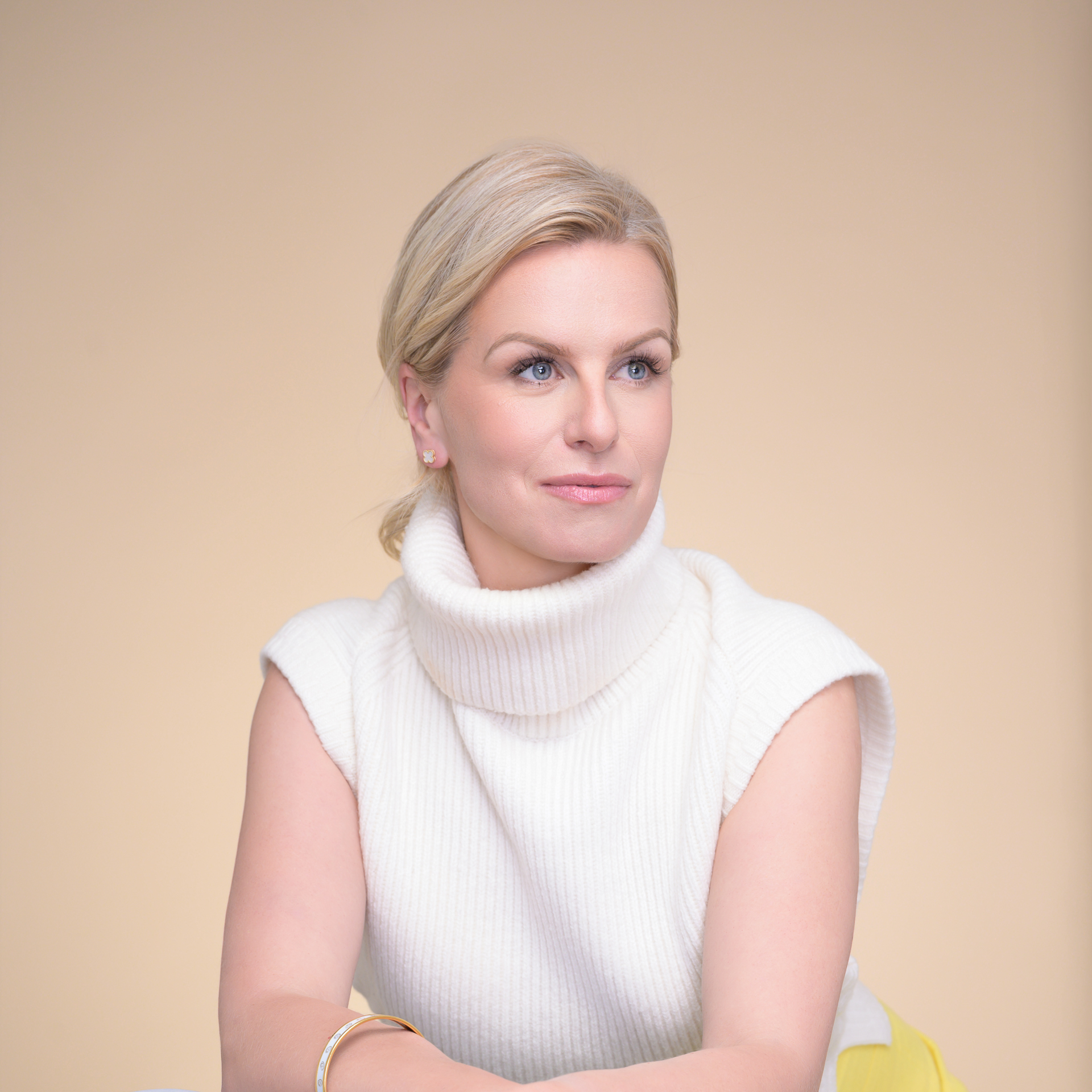 Headshot of Woman with blonde hair wearing a white turtleneck sweater, seated with a neutral background.